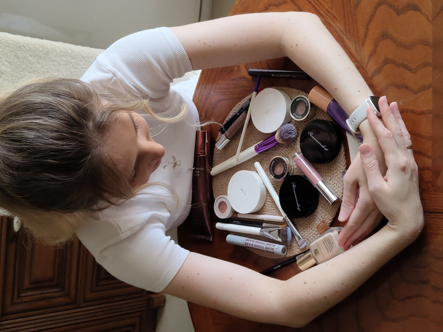 A high-angle, medium shot shows a fair-skinned person with their hands clasped in their lap, looking down at a collection of makeup laid out on a woven mat on a wooden table. The person is wearing a white ribbed t-shirt and has their hair pulled back in a bun with some strands loose around their face. The makeup includes several brushes with purple and white bristles, compacts of various sizes and colors, a tube of foundation, a tube of lip gloss, and a razor. The background is blurred, showing a dark wooden dresser on the left and a light-colored upholstered chair on the right. The image does not provide any information about the location in Krasnodar, Russia.