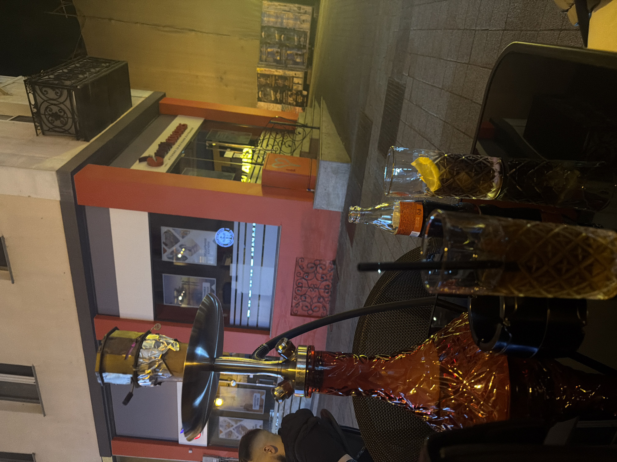 A low-angle shot shows a hookah and two drinks on an outdoor table in Plovdiv, Bulgaria. The table is in the foreground and is made of a dark material with a mesh pattern. The hookah, with a silver and orange base, a bowl wrapped in foil, and a long black hose, sits on the table. Two glasses with straws are next to the hookah. One glass holds a drink with a lemon wedge, and an empty bottle lies next to it. A person is partially visible near the base of the hookah. In the background, the facade of a building with large windows and a red-orange trim is visible. The street and other buildings are further in the background.