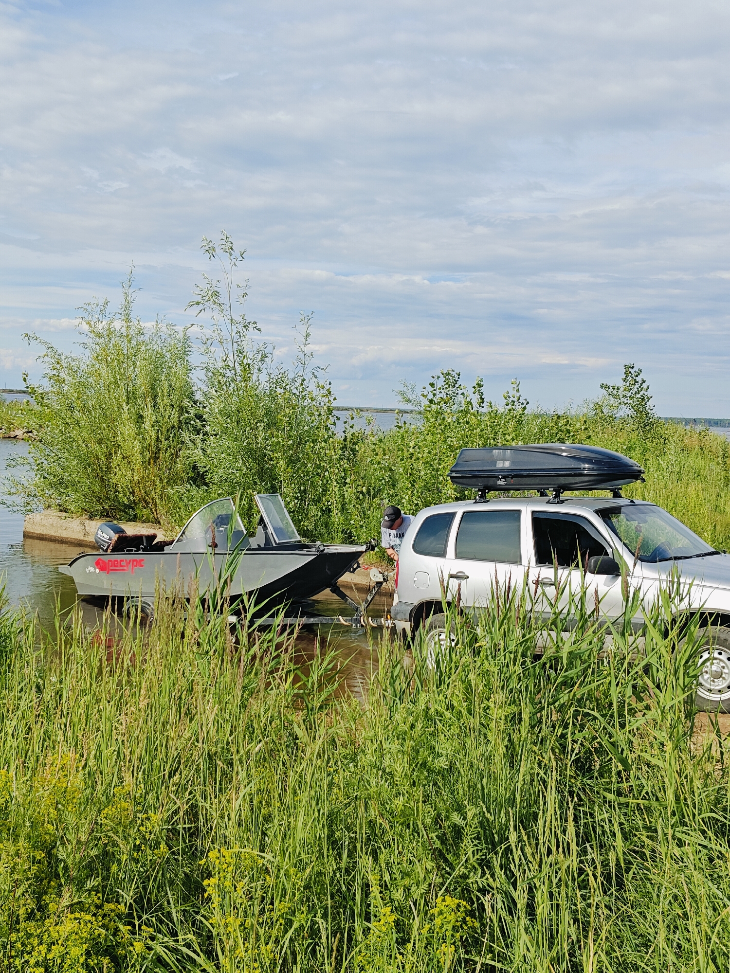 In Chaikovsky, Russia, a silver SUV with a black roof cargo carrier is parked on a grassy embankment next to a body of water. A trailer carrying a grey aluminum motorboat is partially submerged in the water, with the boat's bow resting on the embankment. A person, wearing a black baseball cap and a light-colored t-shirt, is standing between the SUV and the boat, appearing to be engaged with the boat or trailer. Tall green grass and reeds are abundant in the foreground, obscuring the lower parts of the vehicles and obscuring the water's edge. In the background, lush green trees and bushes line the shore, and a calm body of water stretches towards the horizon under a partly cloudy sky. The scene suggests preparations for or the conclusion of a boating activity. The time appears to be daytime, with bright, even lighting indicating it is likely a clear or partly cloudy day. The text "СПЕЦРУС" is visible in red lettering on the side of the boat.