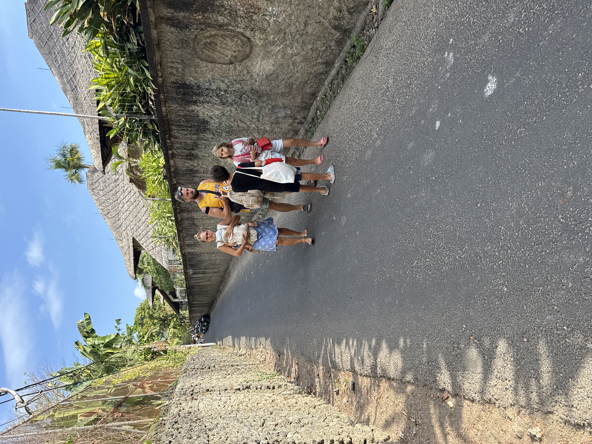 The image depicts a narrow, paved street in Denpasar, Indonesia, lined with a textured stone wall on the left and a concrete wall with tropical vegetation and thatched-roof buildings on the right. The sky is a clear blue with a few wispy clouds. In the foreground, a family of four is walking away from the camera down the street. The father, wearing a yellow and black basketball jersey, walks with a baby strapped to his chest. Beside him, a young girl in a pink shirt and white shorts holds a drink and a red bag. A boy in a black shirt and blue shorts walks in front of the girl, carrying a white bag. Further down the street, several parked motorcycles are visible. The scene suggests a warm, sunny day in a residential or tourist area.
