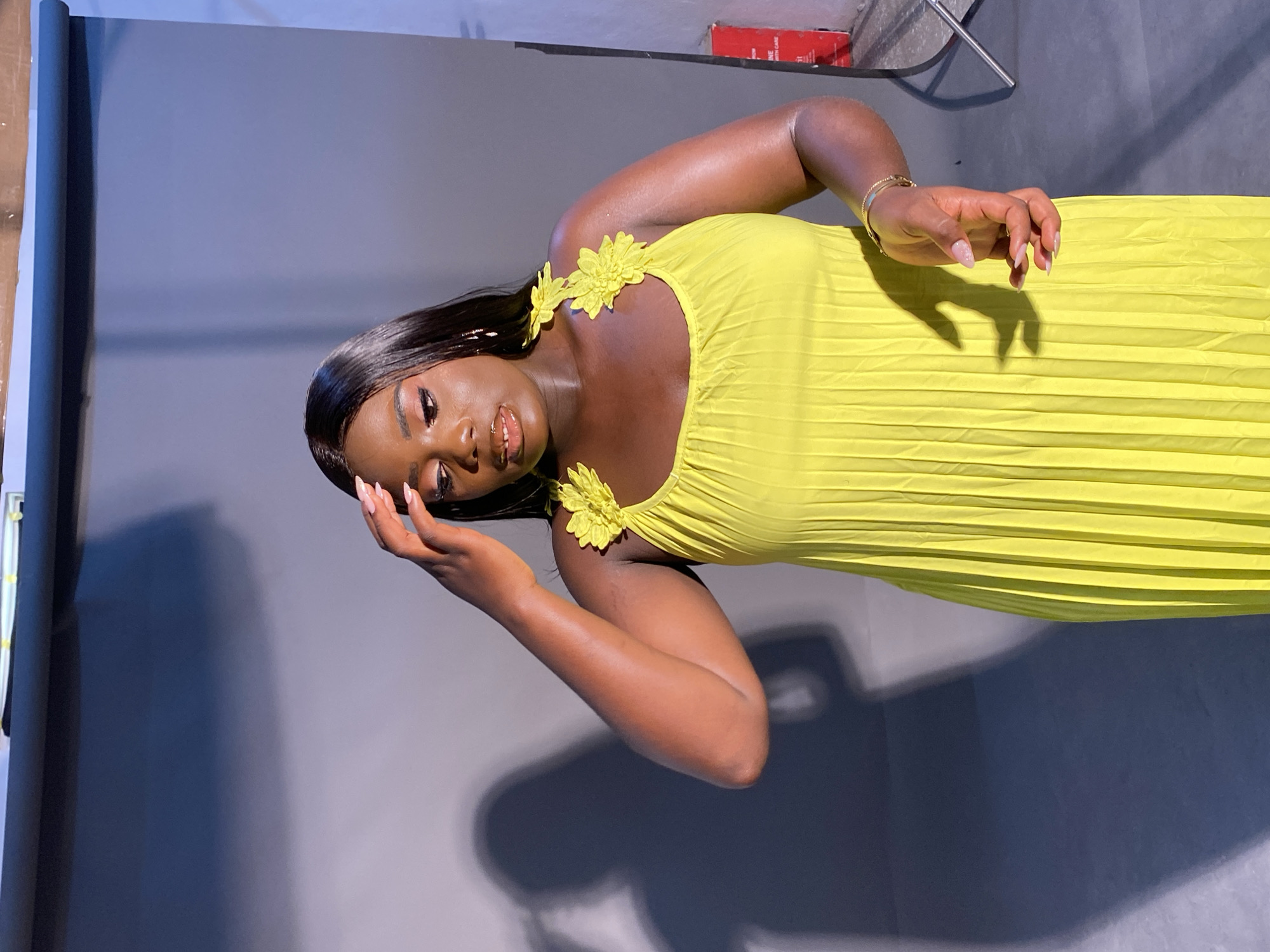 A Black woman is pictured indoors, possibly in a studio setting. She is wearing a bright yellow dress with pleated fabric and decorative floral accents on the shoulder straps. Her right hand is raised to her forehead, and her gaze is directed downwards. Her makeup includes eyeshadow and lip gloss. The background appears to be a plain gray or light blue backdrop with shadows cast by the subject. To the right of the subject, a red box and some metal equipment are partially visible. The lighting creates distinct shadows, suggesting controlled lighting conditions. The location within Gura Top, Nigeria, cannot be determined from the visual information provided in the image.