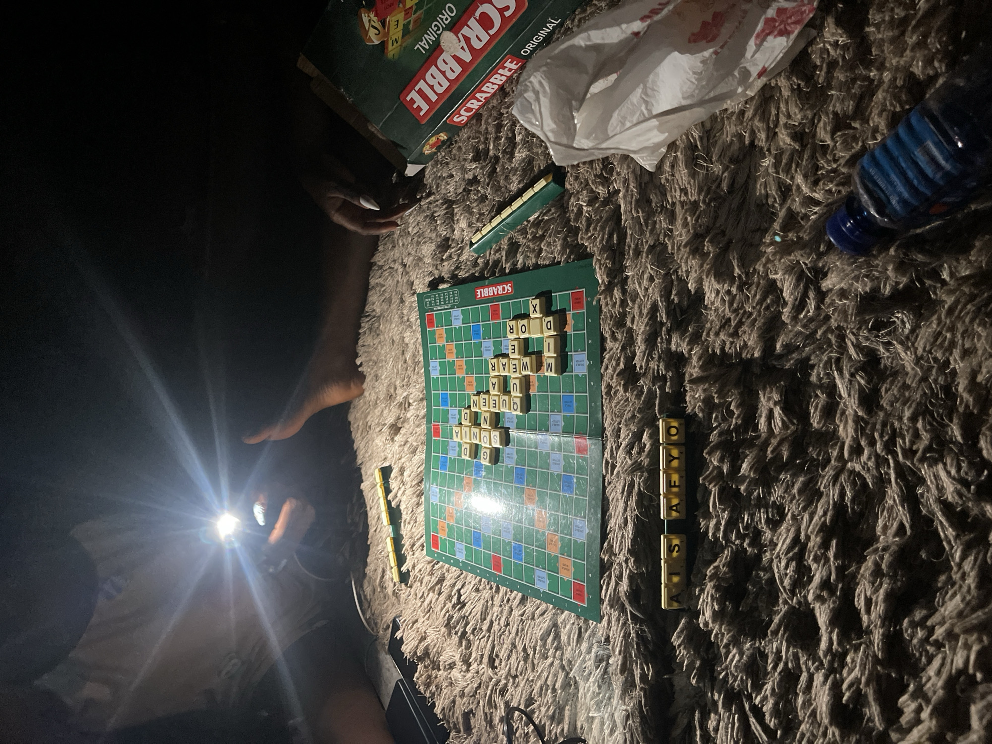 The image depicts a Scrabble game in progress, likely at night or in a dimly lit indoor setting in Kuchingoro, Nigeria. The game is laid out on a shaggy, dark-colored rug. At least two individuals are present, with one person holding a smartphone and using its bright flashlight to illuminate the Scrabble board, creating significant glare. Another person's hand, featuring long white nails, is visible near the "SCRABBLE ORIGINAL" game box.
On the Scrabble board, several words are already formed, including "QUEEN," "WEAR," "SONIA," "DOR," "X," "GD," and "UA." A player's tile rack on the right side of the board holds the letters "ATS AFYO." Another green tile rack is partially visible to the left of the board. Nearby objects include a crumpled white plastic bag and a blue plastic bottle. The primary activity is playing Scrabble, with the strong, focused light from the phone indicating an intentional effort to continue the game despite the low ambient light.