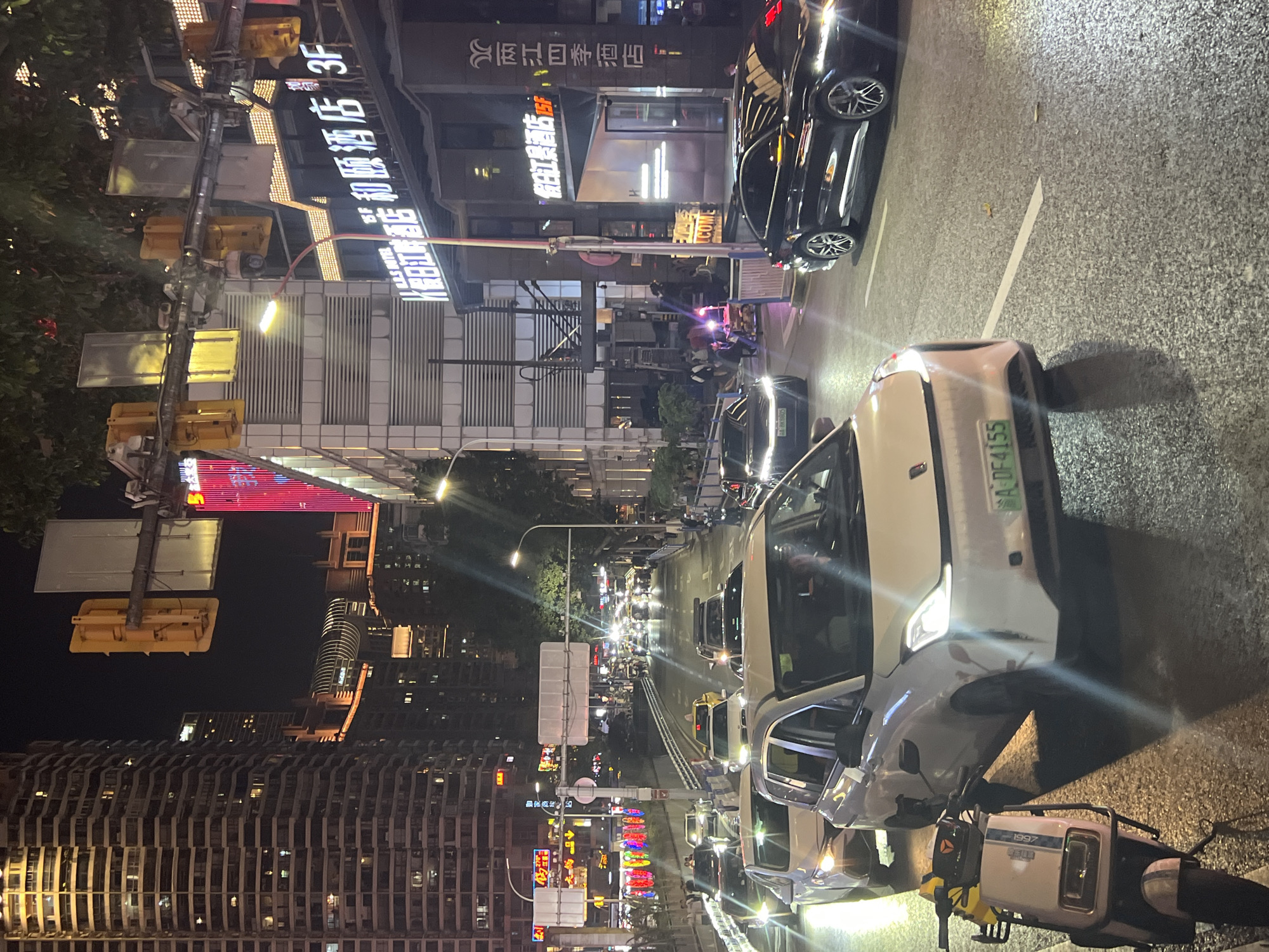 The image captures a nighttime street scene in the Yuzhong District of Chongqing, China, marked by dense urban development. Traffic is a prominent feature with cars lining the street and headlights illuminating the scene. The surrounding buildings are tall and illuminated with neon signs, including what appears to be the "Holiday Inn Riverside Hotel," the "Jihe Yiju Hotel," and the "Rujiang Four Seasons Hotel." Traffic lights and streetlights cast a glow on the area. There are people visible on the sidewalks, though details are indistinct due to the distance and lighting. A distinctive white car with the license plate "A·DF4155" is prominently placed in the foreground, adjacent to a moped.