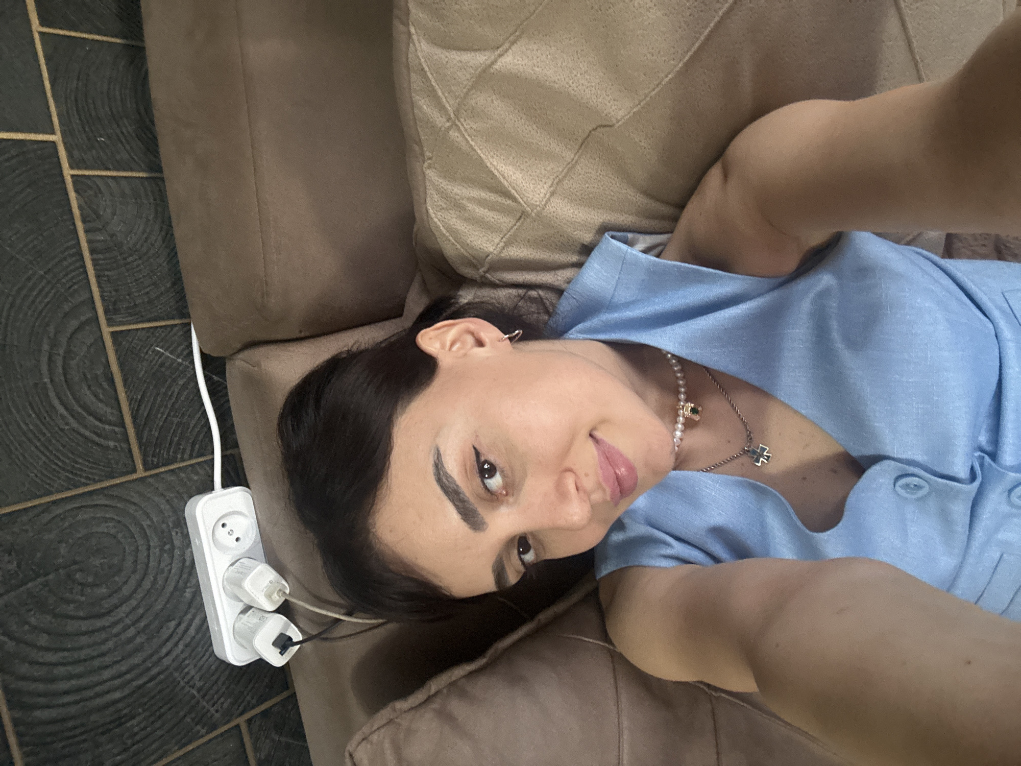 A woman is lying on a brown couch. She is wearing a light blue shirt and a necklace with a cross pendant. The woman has dark hair and fair skin. To the left of the couch is a dark-colored floor. Next to the floor, there is a white power strip with two plugs inserted. The location of the photo is not discernible from the image and cannot be identified as Tbilisi, Georgia.