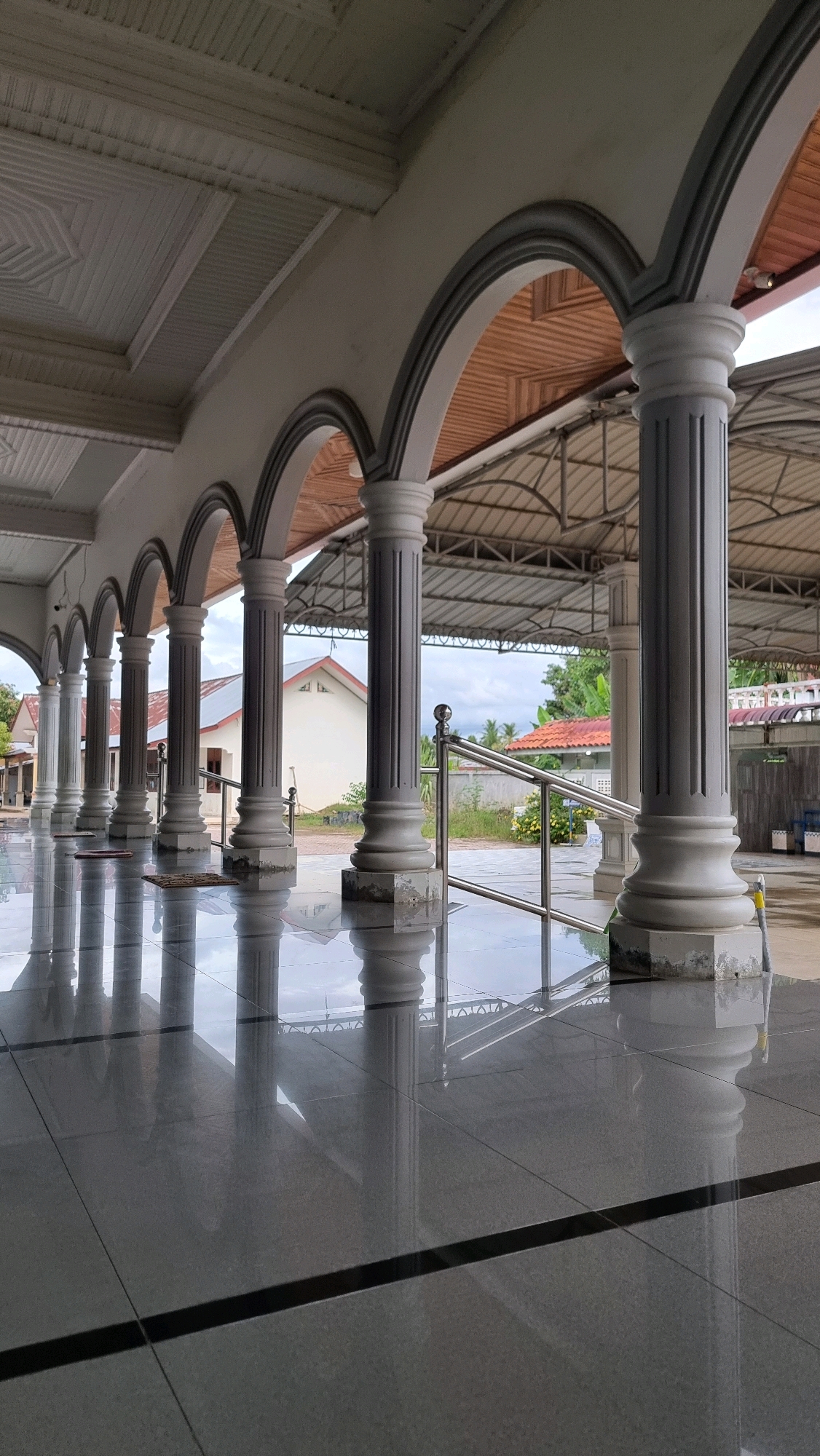 The image shows a covered walkway with a row of fluted columns and arches, leading to an open-air area. The walkway has a polished, reflective tiled floor. In the background, beyond the walkway, are two buildings with red-tiled roofs and a metal railing. Vegetation and a cloudy sky are visible. The architectural style suggests a mosque or a similar public building. The scene is devoid of people. The location is identified as Pantegurah, Indonesia.