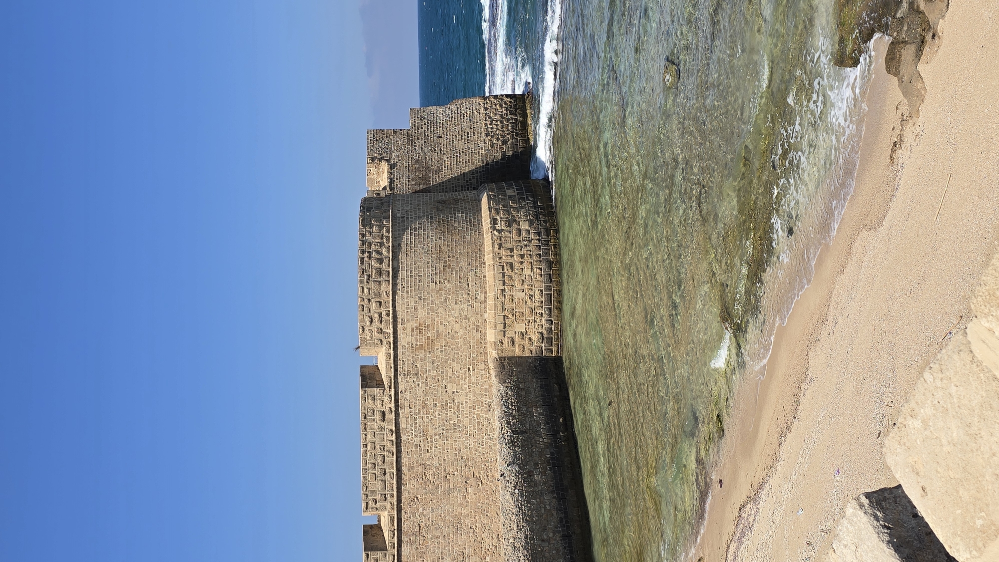 A wide-angle, eye-level shot captures a section of the ancient sea walls of Acre, Israel, under a clear blue sky. The massive stone fortifications, constructed from roughly hewn blocks of light brown stone, dominate the upper two-thirds of the frame. The wall features crenellations along its top edge, with a curved bastion on the left and a more angular, jutting section on the right. Waves gently lap at the base of the wall.
Below the wall is a clear, shallow body of water, revealing the sandy seabed and scattered rocks beneath its surface. The water transitions to a darker, deeper blue further out towards the horizon, where a thin line of clouds is barely visible.
To the left of the water, a sandy beach stretches along the foreground. The sand is light brown and appears fine-grained, with small pebbles and occasional debris. In the extreme lower left corner, a few large, rough-hewn light brown stone blocks create a textured foreground element, suggesting a pathway or steps leading down to the beach. The scene is devoid of people and wildlife, presenting a serene and historic coastal landscape.