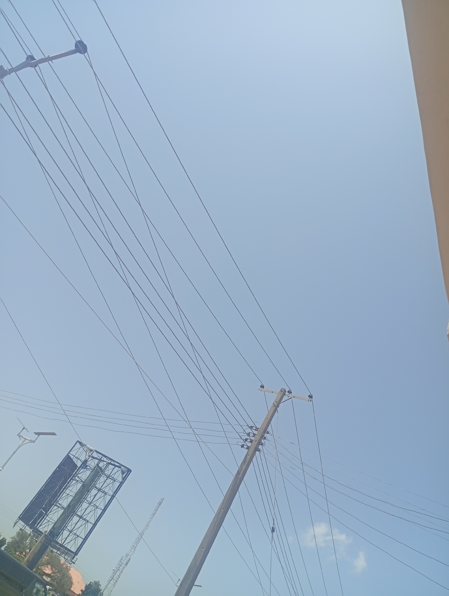 The image shows a bright, clear sky in Bukuru, Nigeria, with numerous utility lines crisscrossing the frame. In the lower left quadrant, a large, empty billboard structure is visible, tilted diagonally. To its left, a street lamp with a curved arm extends upwards. Several wooden utility poles with insulators are scattered throughout the scene, supporting the extensive network of wires. One pole, prominently featured in the mid-right, is angled and supports a dense cluster of wires. In the distance, a tall, lattice-like structure, possibly a communication tower, is discernible. A faint, small cloud is present in the sky to the right of center. The overall impression is one of infrastructure against a vast, open sky, typical of an urban or semi-urban environment in Nigeria.