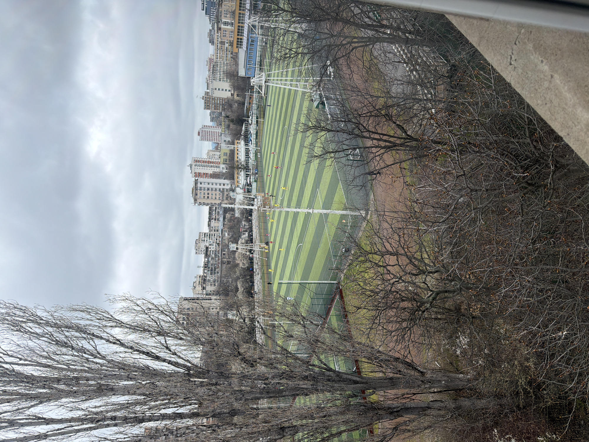 The image shows a soccer field with alternating light and dark green stripes, surrounded by leafless trees. In the background, there are several tall buildings, and the sky is overcast. People in yellow and red jerseys can be seen playing soccer. The location is likely Sofia, Bulgaria.