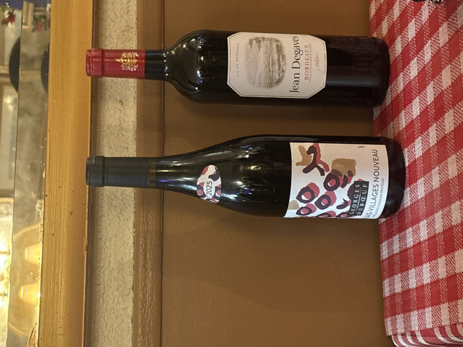 Two wine bottles are positioned horizontally on a brown surface near a red and white checkered cloth. The top bottle, "Jean Degaves Bordeaux 2023," has a red foil neck and a light-colored label. The bottom bottle, "Georges Duboeuf Beaujolais-Villages Nouveau," features a black neck and a white label with abstract shapes and red circles. A small oval sticker with "2025" is affixed to the bottom bottle. The background includes a wooden frame and a reflective surface, likely a mirror.