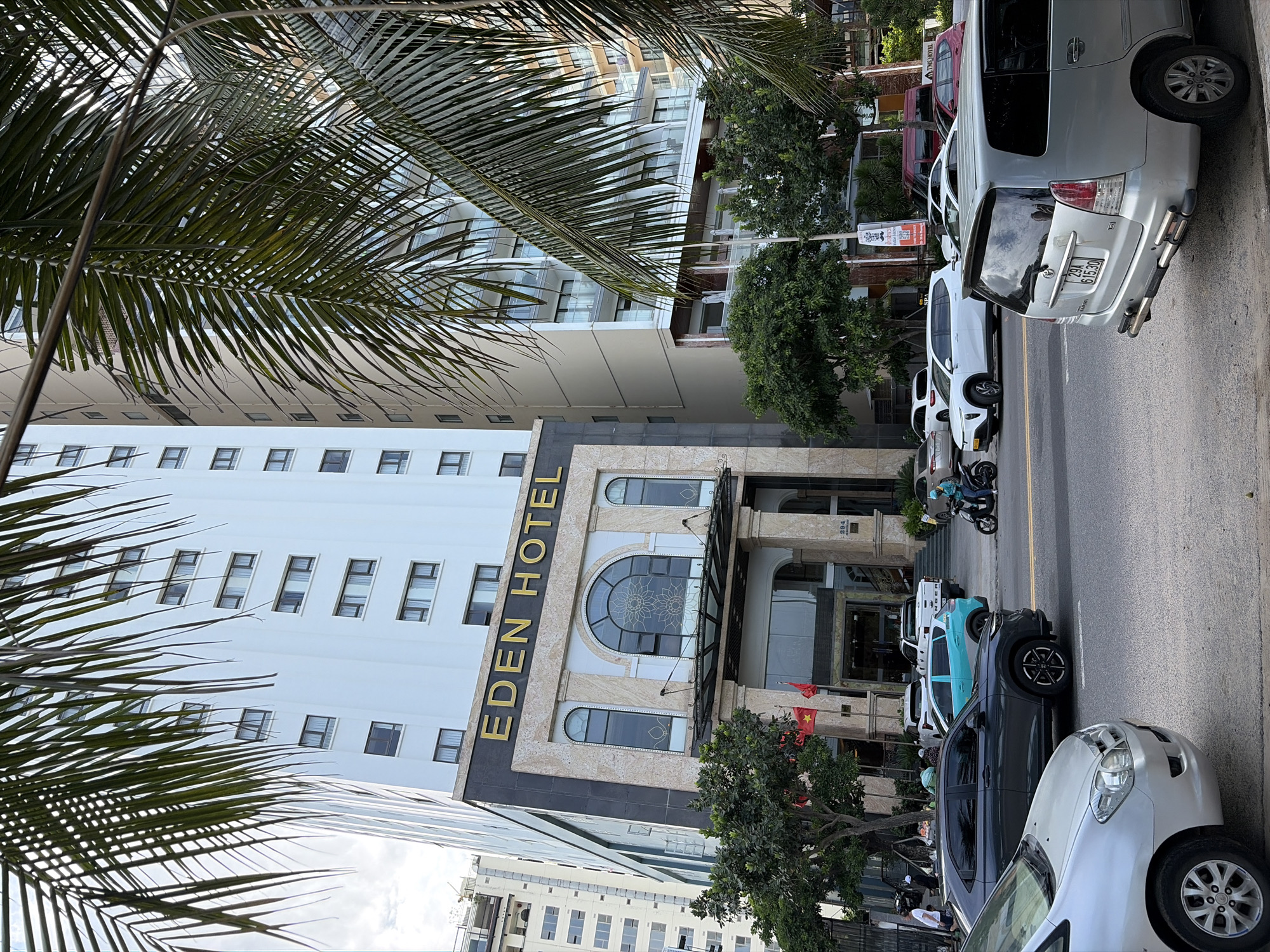 This image captures a street view in front of the Eden Hotel, located at 294, possibly a street number, in the city of Đà Nẵng, Vietnam. The hotel is a tall, multi-story building with a light-colored facade, featuring numerous windows. The entrance to the hotel is grand, with ornate columns and a decorative arched window above the entrance. The "EDEN HOTEL" signage is prominently displayed in gold letters.
In the foreground, a street with asphalt pavement is visible. Several vehicles are parked or driving on the street, including a silver car in the lower left, a dark gray SUV, a light blue hatchback, a white SUV, a silver minivan, and a white pickup truck. A person is seen riding a motorcycle in the middle right of the image.
Palm trees with their fronds arching over the scene are a significant environmental element, suggesting a tropical or subtropical climate. In the background, other buildings of varying heights and architectural styles can be seen, typical of an urban environment. Red and yellow Vietnamese flags are displayed near the hotel entrance. The sky is partly cloudy. The overall impression is a busy street scene in a coastal city.