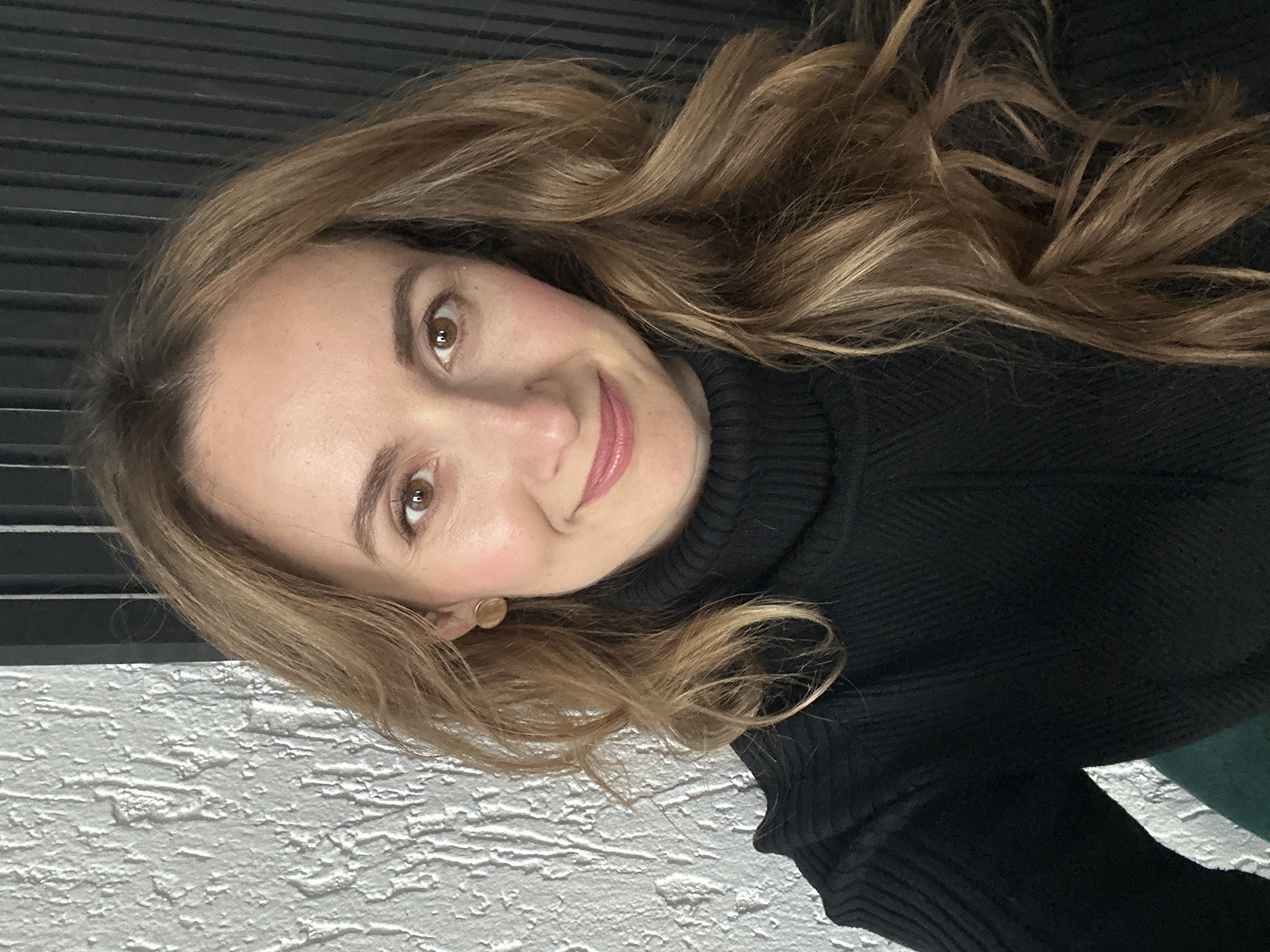 A portrait-style image features a woman with light brown, wavy hair looking directly at the camera with a soft, pleasant smile. Her head is tilted to her left. She has brown eyes, a clear complexion, with a hint of blush on her cheeks and light pink lipstick. Her eyebrows are well-defined.
She is wearing a black turtleneck sweater with a visible textured knit pattern. A small, round, gold-colored stud earring is visible in her right ear.
The background immediately behind her head and shoulders is dark, showing subtle horizontal lines, possibly from blinds or a similar slatted surface. To her lower left, a white, heavily textured wall, resembling stucco or rough plaster, is visible. A small portion of a dark green fabric or object is partially visible in the bottom right corner of the image, behind her right shoulder. The lighting is soft and even, suggesting an indoor setting. There is no visible text in the image. The scene is located in Kryvyi Rih, Ukraine.