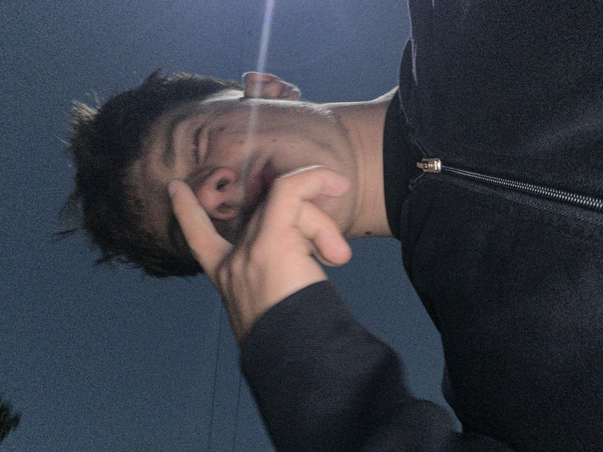 A low-angle, close-up photograph depicts a young male individual in the city of Zlatograd, Bulgaria. The subject is seen from the chest up, wearing a dark, possibly black or navy blue, long-sleeved garment featuring a prominent metallic zipper closure. He has dark, short, disheveled hair and a light amount of facial stubble. His right hand is raised, with the index finger extended towards his forehead or eyebrow area, while the remaining fingers are curled, partially obscuring his nose and mouth. His eyes are partially closed or squinted, and his mouth is slightly open. A distinct horizontal light flare is visible across the right side of his face and ear. The background is a uniform dark blue. The image displays considerable digital noise, consistent with a low-light environment.