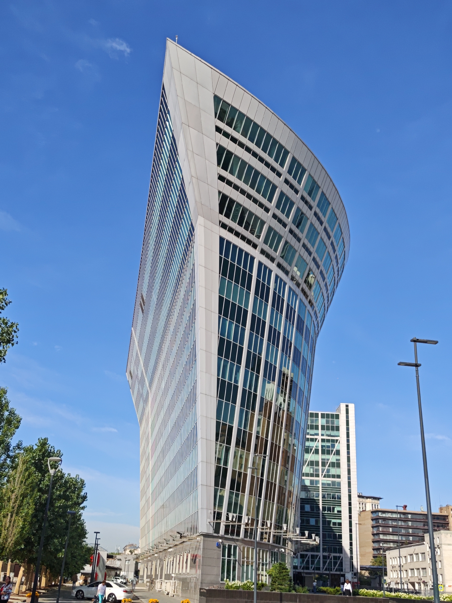 The image depicts the modern architecture of Moscow, Russia, with a prominent focus on the uniquely shaped building known as the Dominion Tower. The Dominion Tower, characterized by its striking curved facade and extensive glass windows, is a contemporary office building located in the Moscow International Business Center. The building's design features a series of stacked, offset floor plates, creating a distinctive, almost triangular profile from certain angles.
In the foreground, the ground level of the Dominion Tower features a transparent canopy or entrance area, with visible architectural details such as support beams and window panes. Several potted plants with orange flowers are placed along the sidewalk, adding a touch of natural color to the urban landscape. A white sedan is parked on the street, and a few individuals are walking on the sidewalk. One woman in a white top and blue pants is near the car.
To the right of the Dominion Tower, a second, taller rectangular building with a green-tinted glass facade is visible. Further in the background, other buildings of varying architectural styles and heights can be seen, indicative of a bustling city environment. A black lamppost with two horizontal lights stands in the mid-right of the image.
The sky is a clear, vibrant blue with a few wispy clouds, suggesting a sunny day. The overall scene conveys a sense of modern urban development and architectural innovation in Moscow.