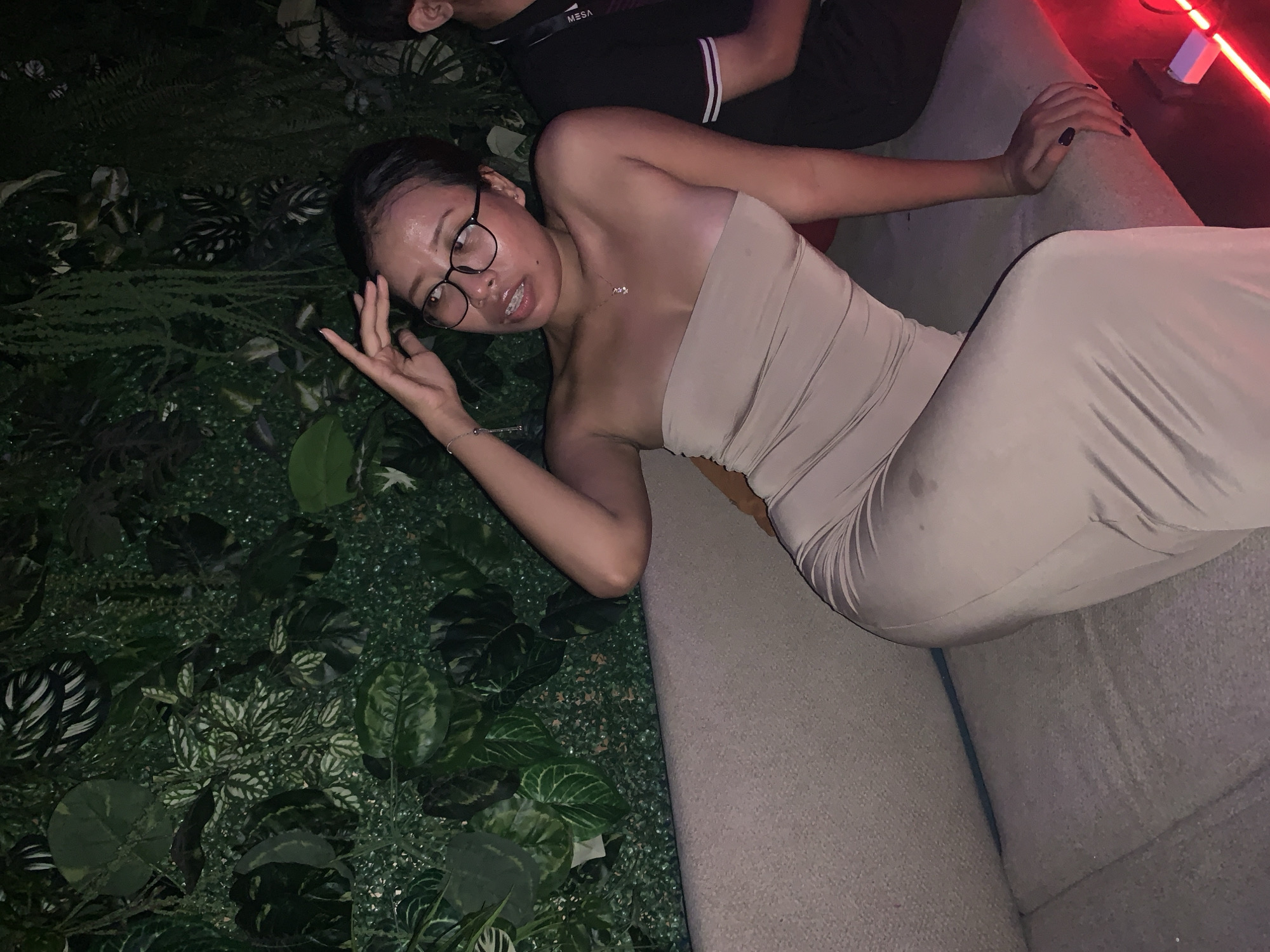 The image shows a young woman with glasses, wearing a strapless beige dress, reclining on a grey sofa. She is making a hand gesture and looking towards the camera. Next to her is another individual partially visible at the top of the frame, wearing a black shirt with white stripes on the sleeves. Part of a faux plant wall is visible on the left side of the frame. A red light source is visible in the top right corner. The location is North Kuta, Indonesia.
