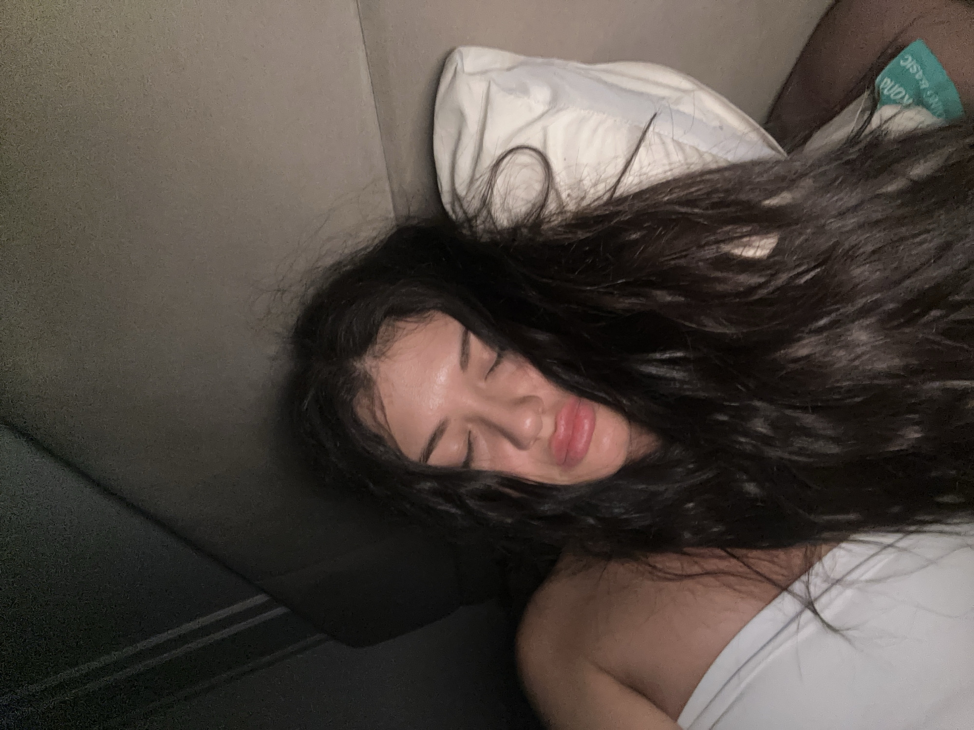 A woman with dark hair is asleep, lying on her side on a grey couch. Her head rests on a white pillow. She is wearing a white top. Part of another person's shoulder and an elastic band with "BASIC" printed on it are visible in the upper right corner. The location is not discernible from the image.