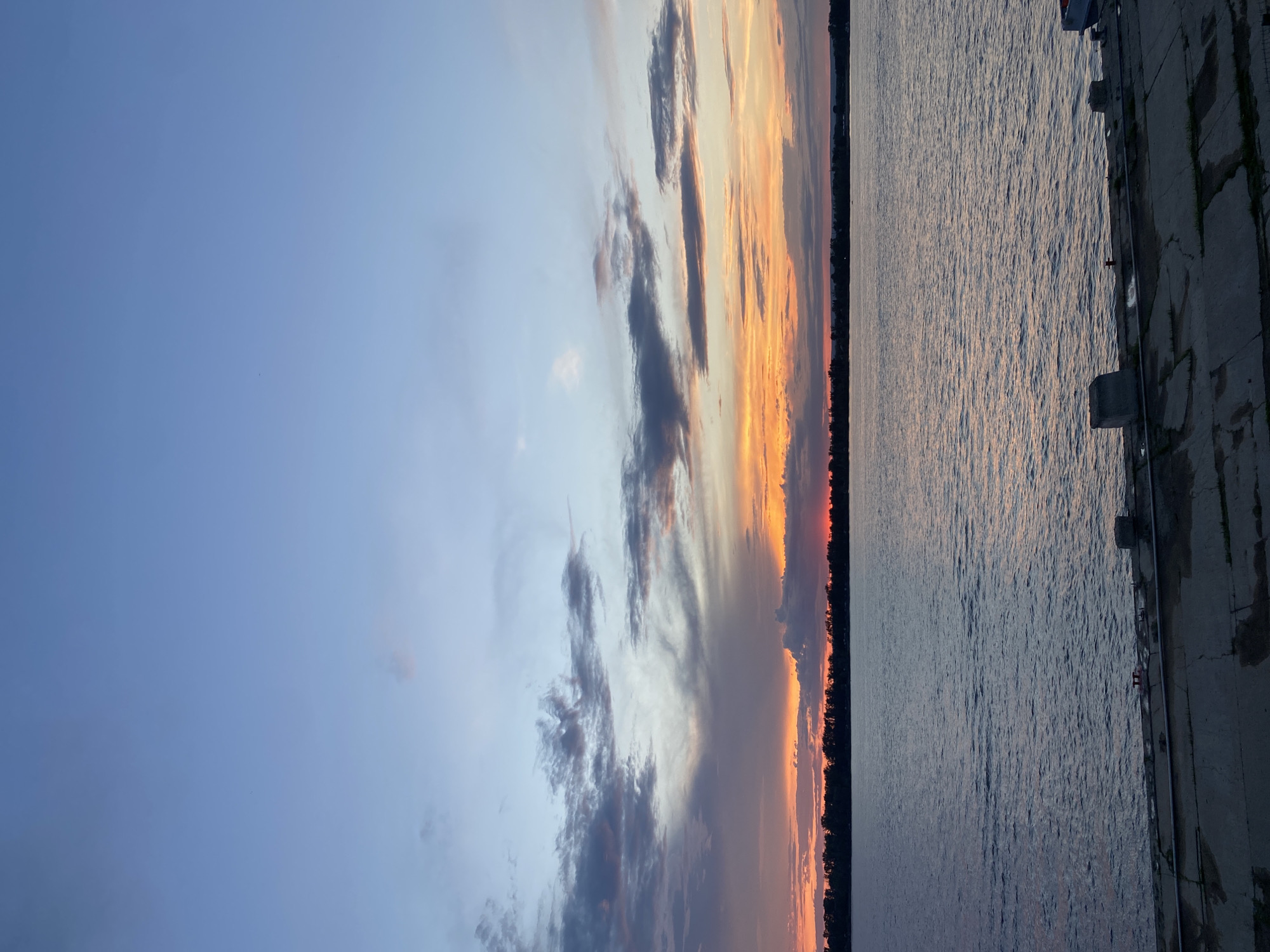 The image depicts a scenic view of the Volga River at sunset, with the cityscape of Novokuybyshevsk, Russia, visible on the horizon. The sky is a gradient of blue and orange, with scattered clouds. The river is calm, with gentle ripples reflecting the colors of the sky. In the foreground, there is a concrete embankment with several bollards and some debris. The scene is tranquil and serene, capturing the beauty of a Russian sunset.