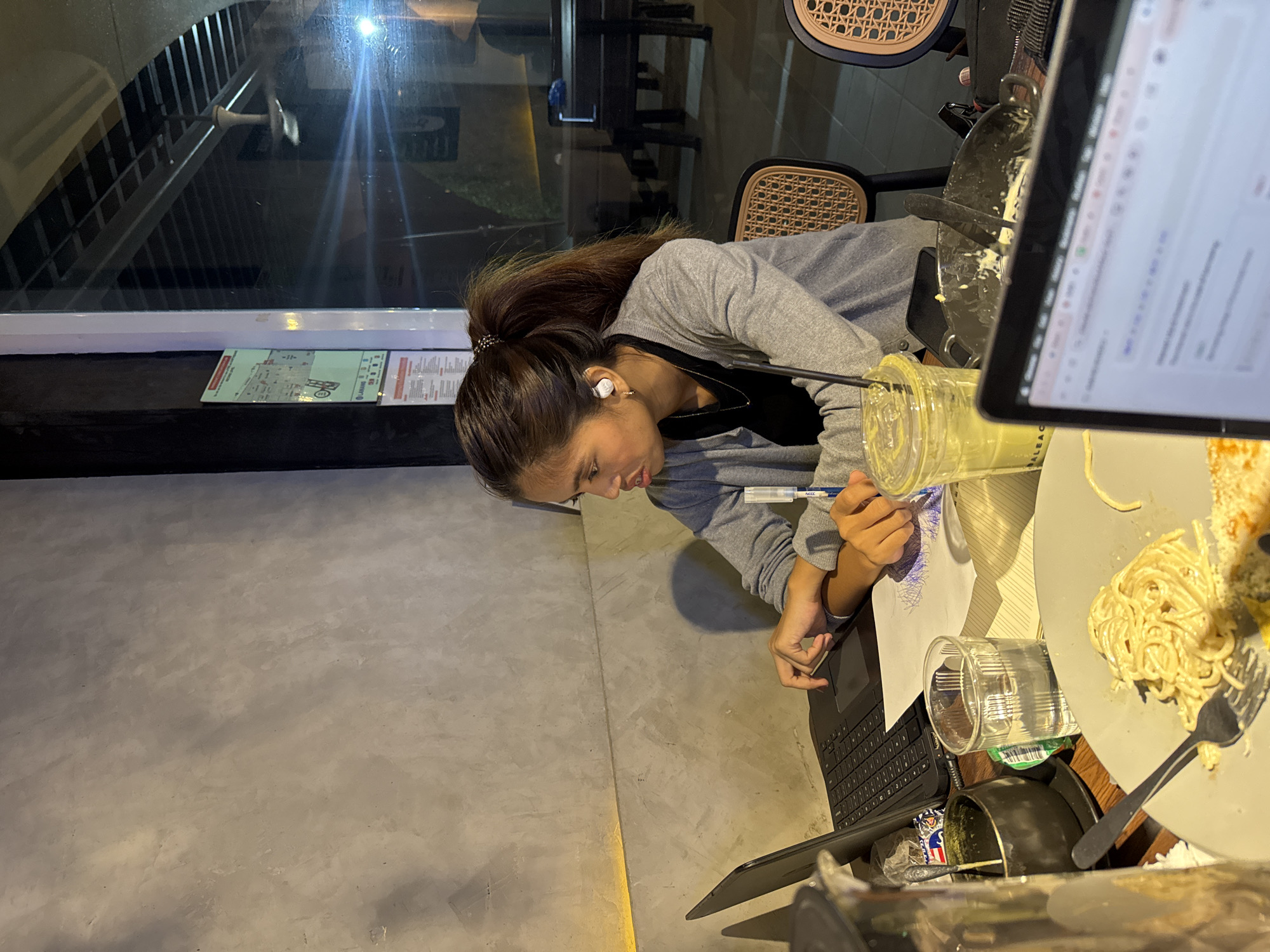 A person is bent over a table, drawing on a piece of paper. The person has long brown hair pulled back in a ponytail and is wearing a gray sweater. An earbud is visible in their left ear. They are holding a pen and looking intently at the paper.
The table is cluttered with various items, including a laptop, a glass of clear liquid, a smoothie, and a plate of pasta. A fork is resting on the plate. The table is next to a gray concrete wall.
Outside the window, there is a view of a building with outdoor stairs. The lighting suggests it is evening or night.
Based on a sign visible in the image ("Labang"), this is likely a location in Davao City, Philippines.