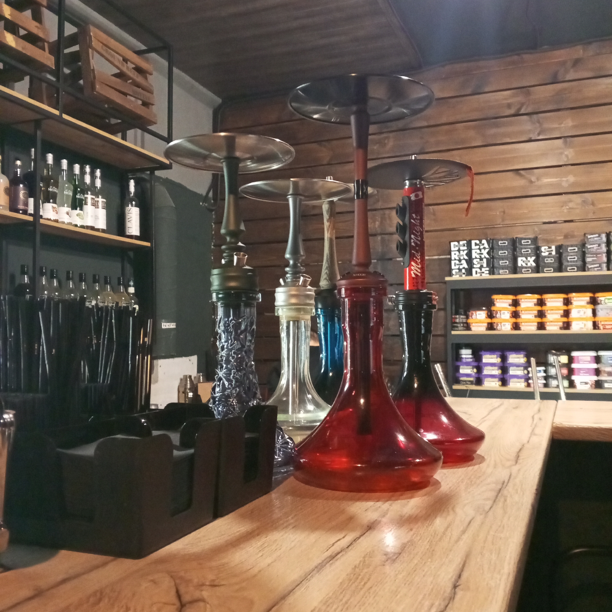 The image shows a row of hookahs on a wooden counter, likely in a bar or lounge. The hookahs are various colors, including blue, clear, and red. Behind the counter are shelves stocked with liquor bottles, hookah tobacco, and other supplies. The location is likely in the city of Chaikovsky, Russia.