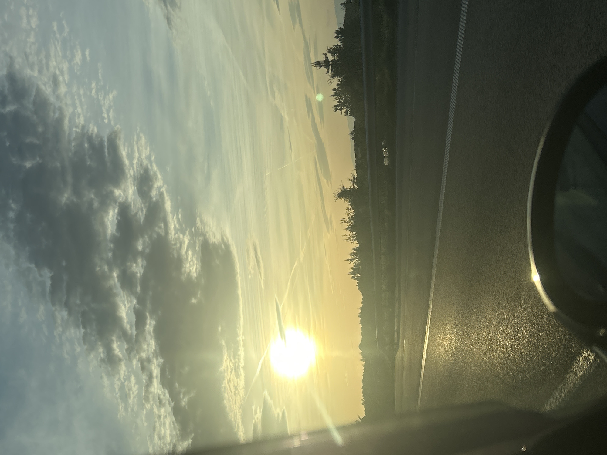 The image shows a view from inside a vehicle, likely a car, during a sunset or sunrise. The sky is filled with clouds on the left side, transitioning to a brighter, yellowish hue towards the center where the sun is positioned. The sun is low on the horizon, creating a bright glare. A road or highway runs parallel on the right side of the image, and a line of trees or foliage is visible along the road's edge. The interior of the car is partially visible, with the side mirror appearing in the bottom right corner. The location appears to be an open area with no specific city or landmark apparent.