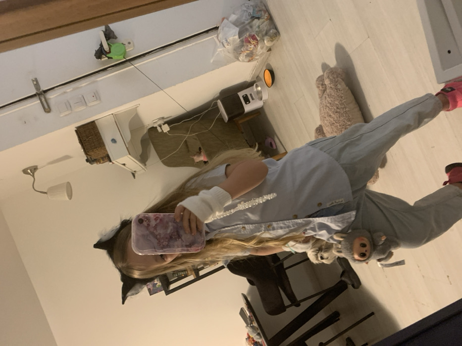 The image, rotated 90 degrees counter-clockwise, depicts a young person taking a mirror selfie indoors. The primary subject has long blonde hair, which falls past their shoulders. They are wearing a light blue button-up shirt, light-colored long pants, and pink athletic shoes. A white fingerless arm warmer is visible on their left arm, and they are sporting a black cat ear headband.
The person is holding a smartphone with a distinctive purple and pink marble-patterned case, which largely obscures their face as they capture the image. Attached to their hip or pants is a small, gray plush toy resembling a monkey.
The reflected background reveals an indoor setting, likely a room in a home with white walls and a light-colored floor, possibly wood or laminate. Visible objects include a white cabinet or small dresser with a woven basket on top, a wall-mounted lamp with a white shade, and what appears to be a projector or humidifier situated on a small table. Other details include electrical outlets or switches on the wall, a green and white device attached to the wall (possibly a retractable clothesline or toy), and several plastic bags containing various items, contributing to a somewhat casual, lived-in atmosphere.
The lighting suggests an indoor setting with artificial light, not strong daylight. No discernible text is visible in the image. The provided context indicates the location is Bat Yam, Israel.