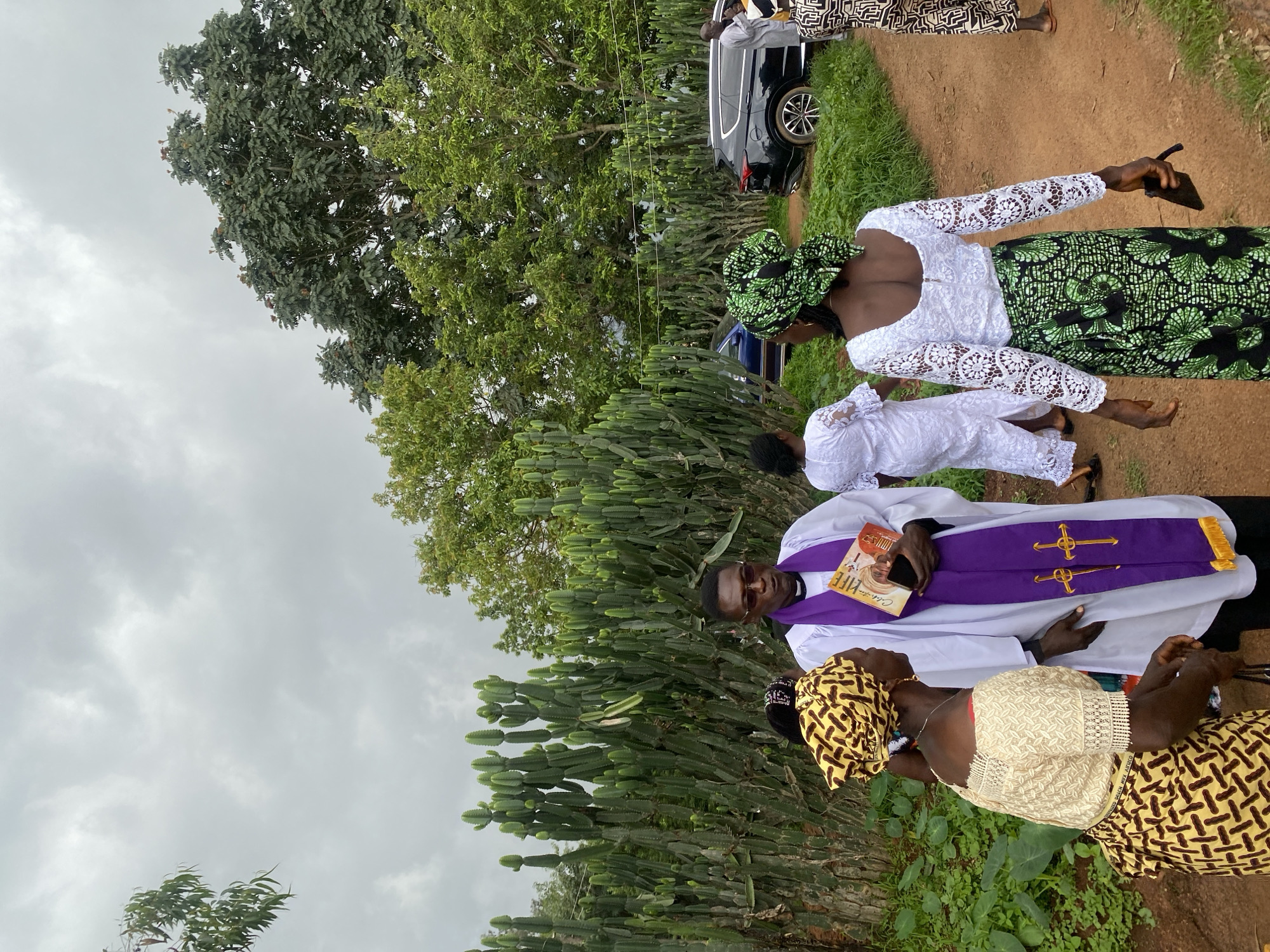 The image depicts an outdoor gathering in what appears to be a rural setting near Wang, Nigeria. A person dressed in clerical attire, a white robe with a purple stole adorned with gold crosses, stands holding a booklet. They are conversing with two women, one wearing a yellow headwrap and a cream-colored lace top with a patterned skirt, and another wearing a green headwrap and a white lace top with a green and black patterned skirt. In the background, a dense hedge of tall, segmented cacti lines a dirt path. Beyond the cacti, lush green trees are visible, and a black SUV and a blue sedan are parked. The sky is overcast with gray clouds. The scene suggests a social event or religious gathering taking place outdoors in a Nigerian locale.