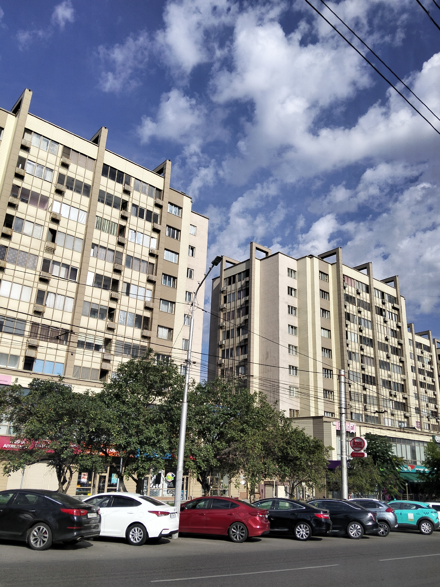 The image captures a street scene in Almaty, Kazakhstan, under a partly cloudy blue sky. The foreground shows a row of parked cars on an asphalt road, including a black sedan, a white sedan, a red sedan, and other vehicles further down the street. Behind the cars, a line of trees with lush green foliage separates the road from a row of buildings. The buildings are tall, multi-story structures with a predominantly beige and light brown facade, featuring numerous windows and visible air conditioning units. Power lines crisscross the scene, strung between poles. In the background, more of the same architecture is visible, suggesting a densely built urban environment. The overall impression is a typical city street in Almaty on a bright day.