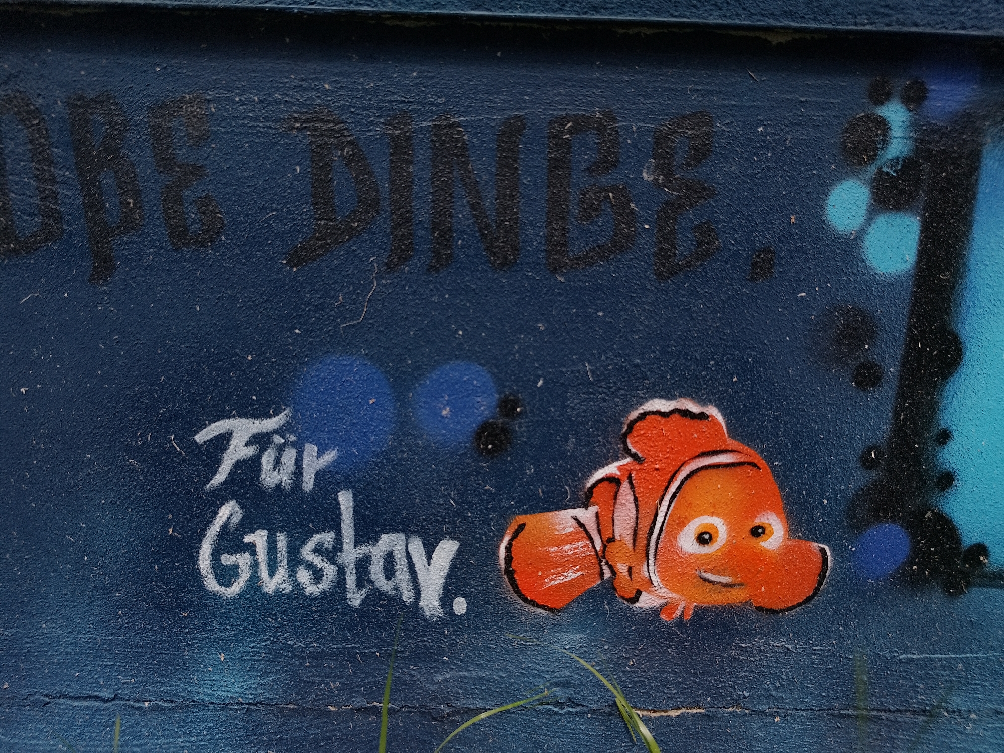 On a rough blue surface, possibly a wall in Unknown, Unknown, graffiti art displays the partially visible German phrase "DIE DINGE." Below this, the phrase "Für Gustav." is written in white paint. An orange clownfish cartoon character is stenciled next to the phrase. Adjacent to the fish are black and blue stenciled shapes. Blades of grass are at the bottom edge of the frame.