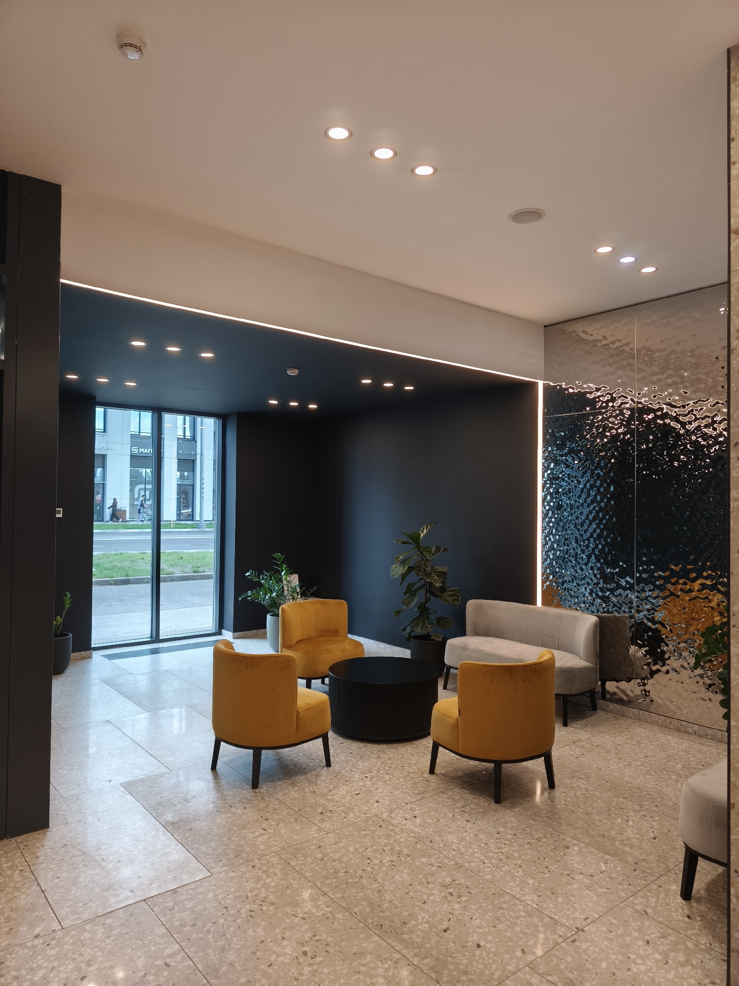 The image captures the interior of a modern lobby or waiting area in Moscow, Russia. The scene is well-lit by recessed ceiling lights and a linear light strip, illuminating the terrazzo floor. A large window on the left reveals a view of a street with pedestrians and a building with the signage "MATI" on it.
Furniture consists of three mustard-yellow armchairs arranged around a round black coffee table. A light grey loveseat and another armchair are positioned against a wall covered in a reflective, textured silver material. Potted plants are placed around the room, adding a touch of greenery. The walls are dark grey, and the space appears clean and well-maintained. There are no people in the immediate foreground of the photo, but figures are visible outside the window.