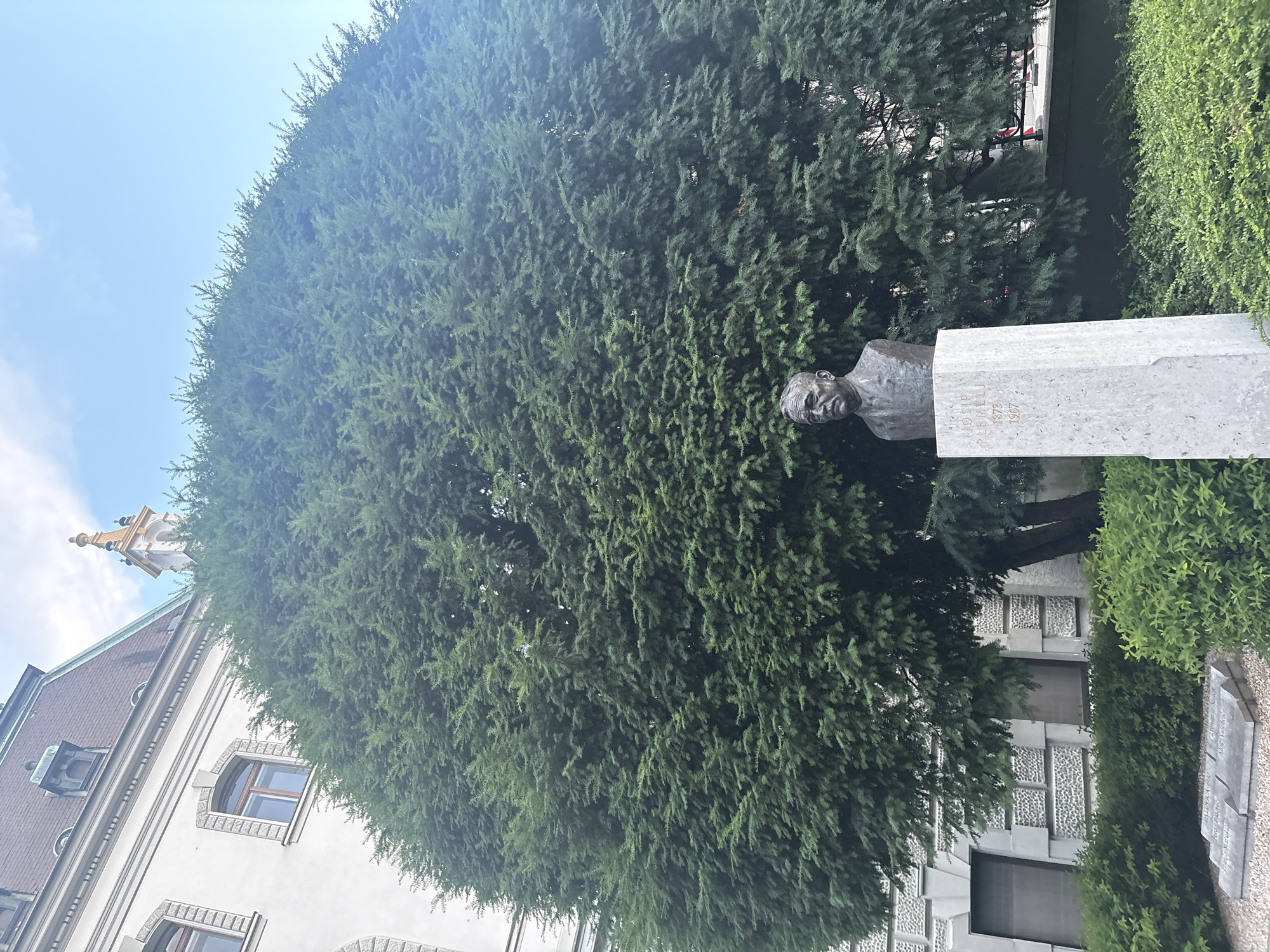 A bronze bust of a man with a mustache and a suit stands atop a white stone monument in front of a large, dense evergreen tree in a courtyard. The monument has text inscribed on it that reads "JOSIP" in the top line, "PLEMELJ" in the second line, and "1873" and "1967" on the third and fourth lines respectively. The man in the bust appears to be of middle age. Behind the tree, a white building with arched windows and a dark, pitched roof is visible. The sky is blue with some white clouds. In the foreground, there are manicured green bushes. To the left of the monument, there are two flat gravestones with inscribed text, some of which is obscured by shadow and blur. The overall scene suggests a memorial or monument located in an urban setting.