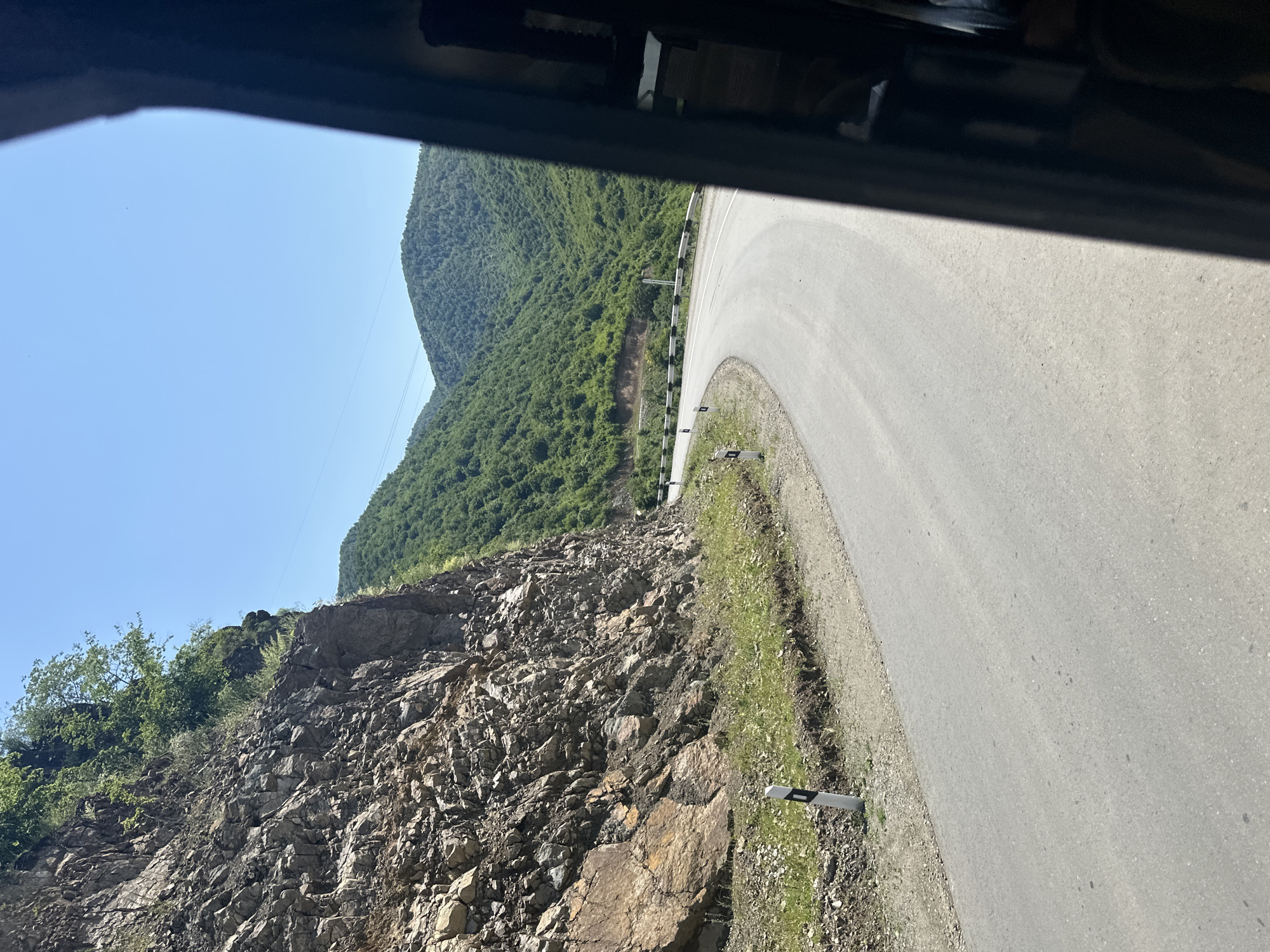 The image depicts a winding road on a sunny day, carved into a mountainous landscape. On the left side of the road is a steep, rocky cliff face covered in sparse vegetation. The road itself is paved and appears to be in good condition, with a guardrail visible along the outside of a curve. Lush green, forested mountains rise on the right side of the road, stretching into the distance. The sky is clear and blue. The view is captured from inside a vehicle, with the dark interior frame visible along the right edge of the image. This scene is consistent with the mountainous terrain surrounding Alaverdi, Armenia, known for its scenic roads and natural beauty.