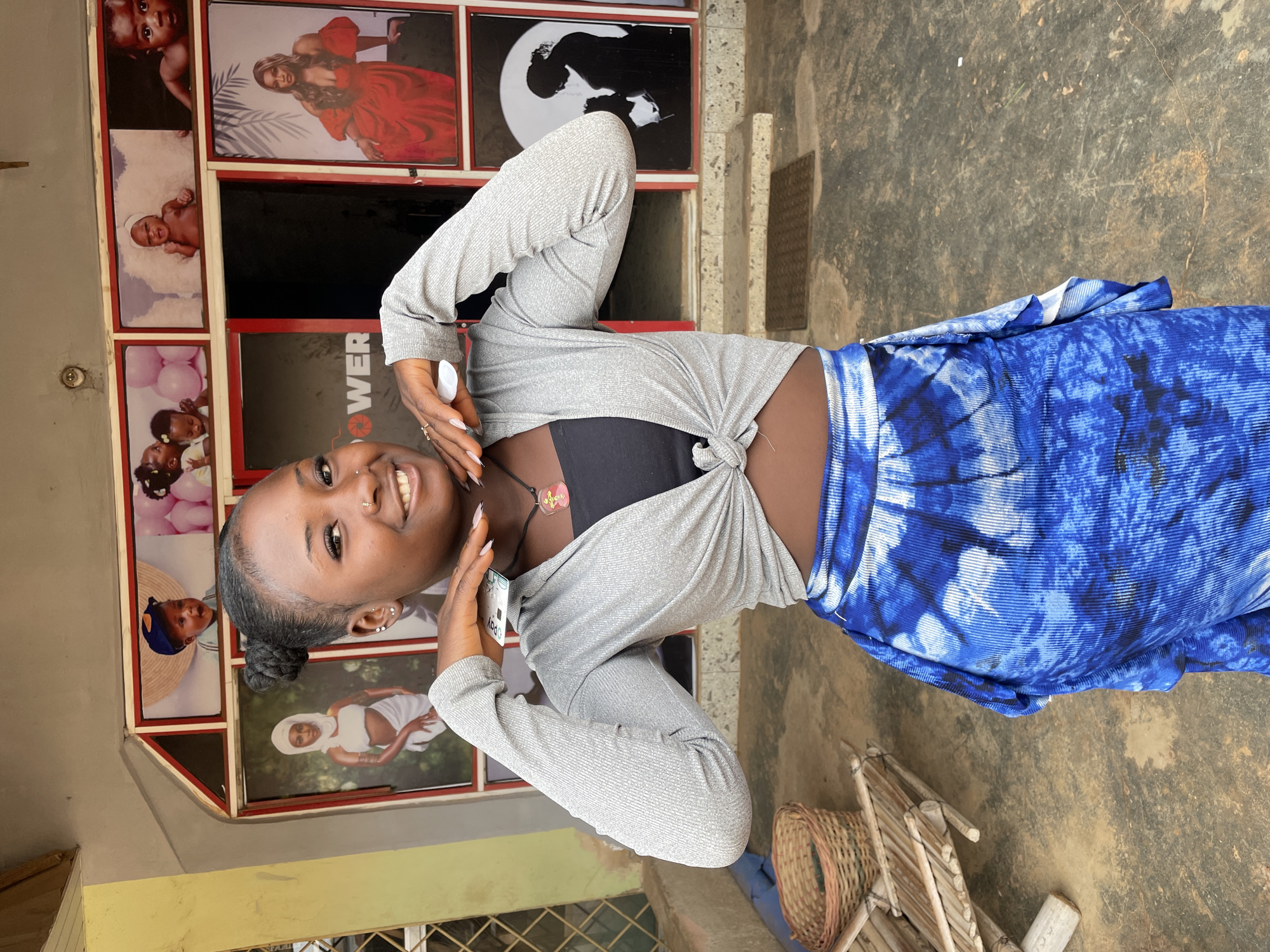 A young woman stands and smiles in front of a building exterior. The building has a display of photographs of people and babies in a grid pattern. The woman is wearing a gray knit top tied at the waist, a black top underneath, and a blue tie-dye skirt or wrap. She is holding an object to her face near her right ear, possibly a phone. The ground appears to be concrete or paved, with a basket and some wooden construction material visible near the bottom right corner. The photograph was taken in Barakin Akawo, Nigeria.