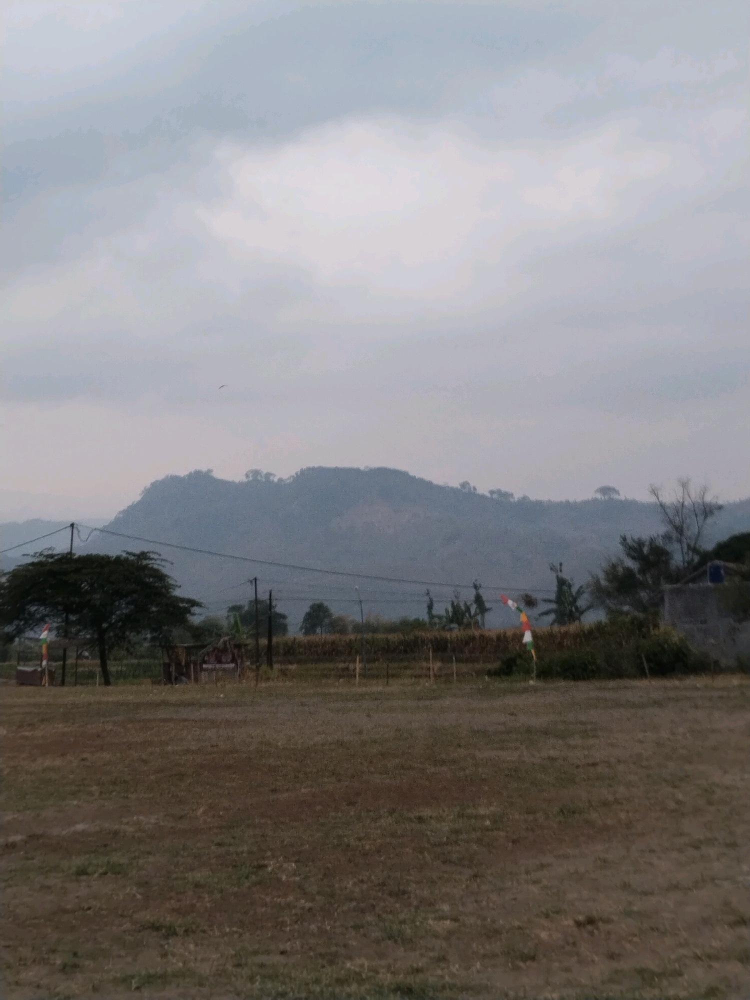The image depicts a landscape scene with a mountain range in the background, under a cloudy sky. In the middle ground, there is a large tree on the left, and some structures and vegetation. A tall pole with a red and white flag, possibly the Indonesian flag, is visible on the right side of the middle ground. The foreground consists of dry, grassy ground. The image appears to be taken in an outdoor setting. The location is Jembayat, Indonesia.