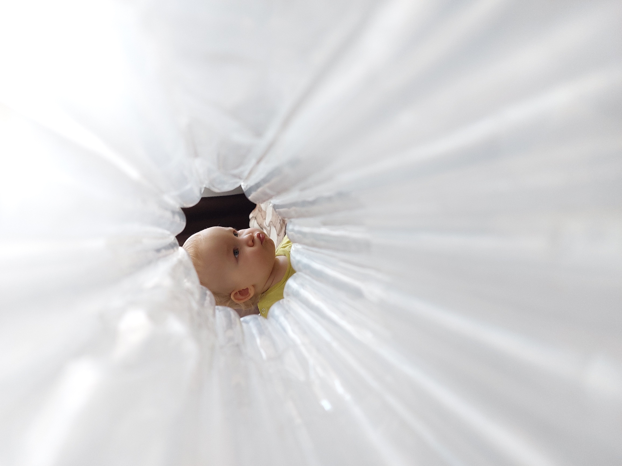 A close-up shot captures an infant partially framed by a semi-transparent white material. The infant has fair skin, blonde hair, and is wearing a yellow shirt. The child is looking towards the upper-left of the frame. The photo appears to have been taken in the city of Chaikovsky, Russia.
