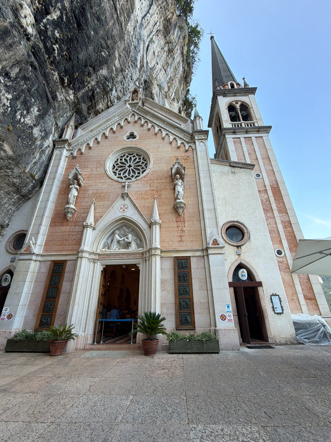 The image shows the Santuario della Madonna della Corona, a church in Ferrara di Monte Baldo, Italy, built into a cliff face. The church has a tall, narrow bell tower with arched openings for bells, and a conical spire. The facade of the church is made of pink and white stone, with a large rose window above the entrance. There are statues of saints on either side of the entrance. The entrance is open, and people can be seen inside. The church is surrounded by a stone plaza, with potted plants on either side of the entrance. A white umbrella is set up to the right of the church, and a tarp-covered object is visible beyond that. The cliff face behind the church is rough and rocky, with some vegetation growing at the top. The sky is clear and blue.