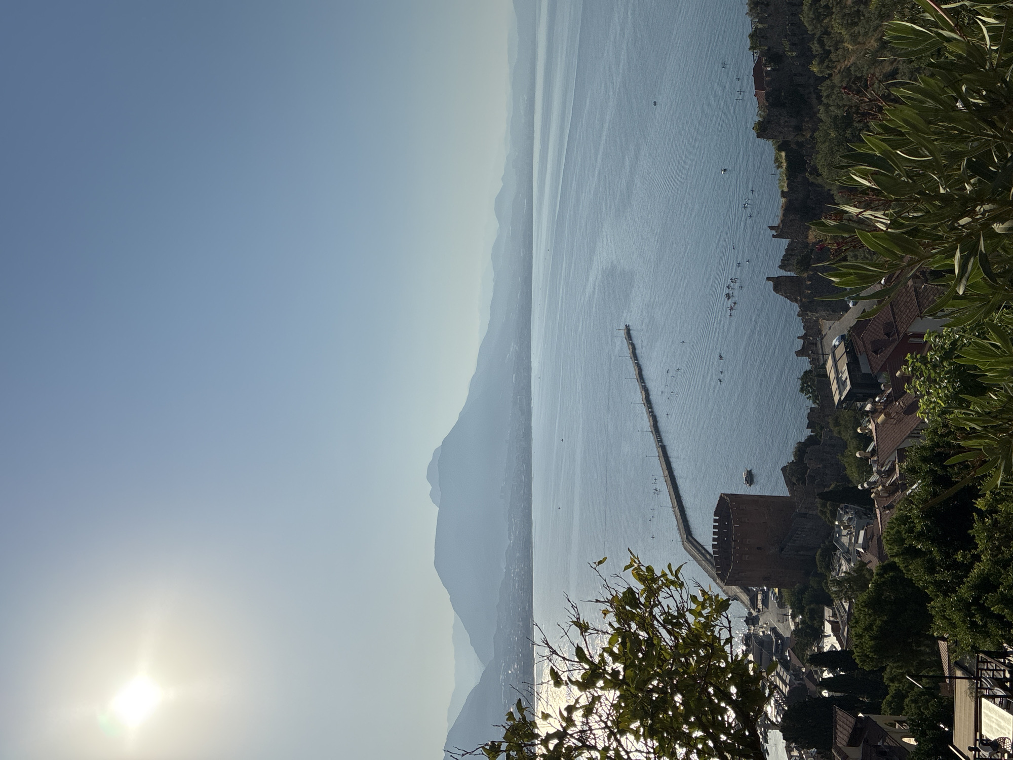 This aerial view shows a bright, sunny day in Alanya, Turkey, with the sun at the upper left quadrant of the image. The clear blue sky transitions to a hazy horizon where distant mountains meet the sea. The water is a deep blue with gentle ripples, reflecting the sunlight. A long pier extends from the coast into the sea, with numerous small figures, likely people, visible along its length and in the water around it.
In the foreground, on the left, lush green foliage partially obscures the view. Below the pier, a prominent, circular, reddish-brown tower stands on the coastline, likely part of the Alanya Castle complex. The tower is surrounded by ancient stone walls and a cluster of buildings with terracotta roofs, indicative of the city's architecture. The coastline stretches out, with more buildings and greenery dotting the landscape. The overall scene captures a vibrant coastal city on a clear, warm day.