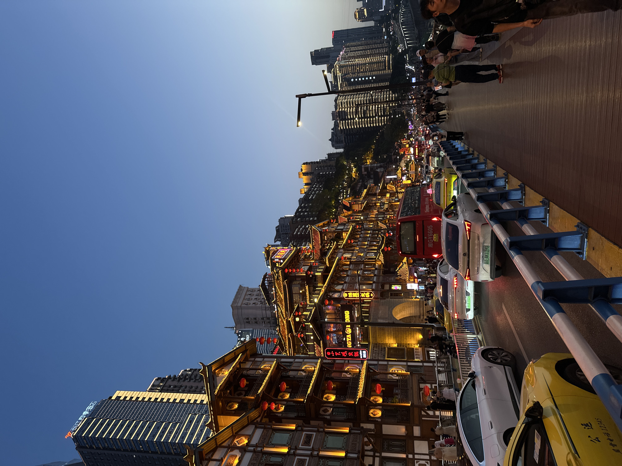 This vibrant night scene, captured in 江北城街道, China, presents a bustling urban landscape at dusk. The sky overhead transitions from a deep blue to a lighter hue towards the horizon, indicating the fading light of day.
The foreground features a wide boardwalk-style sidewalk, where numerous pedestrians stroll and congregate. A blue and white barrier separates this pedestrian area from a busy road to the left, which is filled with vehicular traffic, including several cars, a red bus, and a yellow taxi. Beyond the road, a dense array of multi-story buildings, characterized by traditional Chinese architecture with ornate roofs, wooden facades, and an abundance of warm yellow and red lighting, line the street. These structures are adorned with glowing signs and red lanterns, creating a lively atmosphere. In the background, modern high-rise buildings with illuminated windows punctuate the city skyline, contrasting with the traditional architecture below.
Visible activities include people walking along the boardwalk and street, likely enjoying an evening out, dining, or shopping. The presence of numerous vehicles suggests active commerce and transit. The overall impression is one of a vibrant, energetic urban environment.
Notable visible text includes:
* On the yellow taxi: "公运" (Gōngyùn) and "监督电话 12345" (Supervision hotline 12345).
* On car license plates: "A·879559" and "A·K1390".
* On the red bus: "Shenglong 18 travel" and "湘G-Y6605".
* Partially legible text on building signs such as "NGYAI ONG".