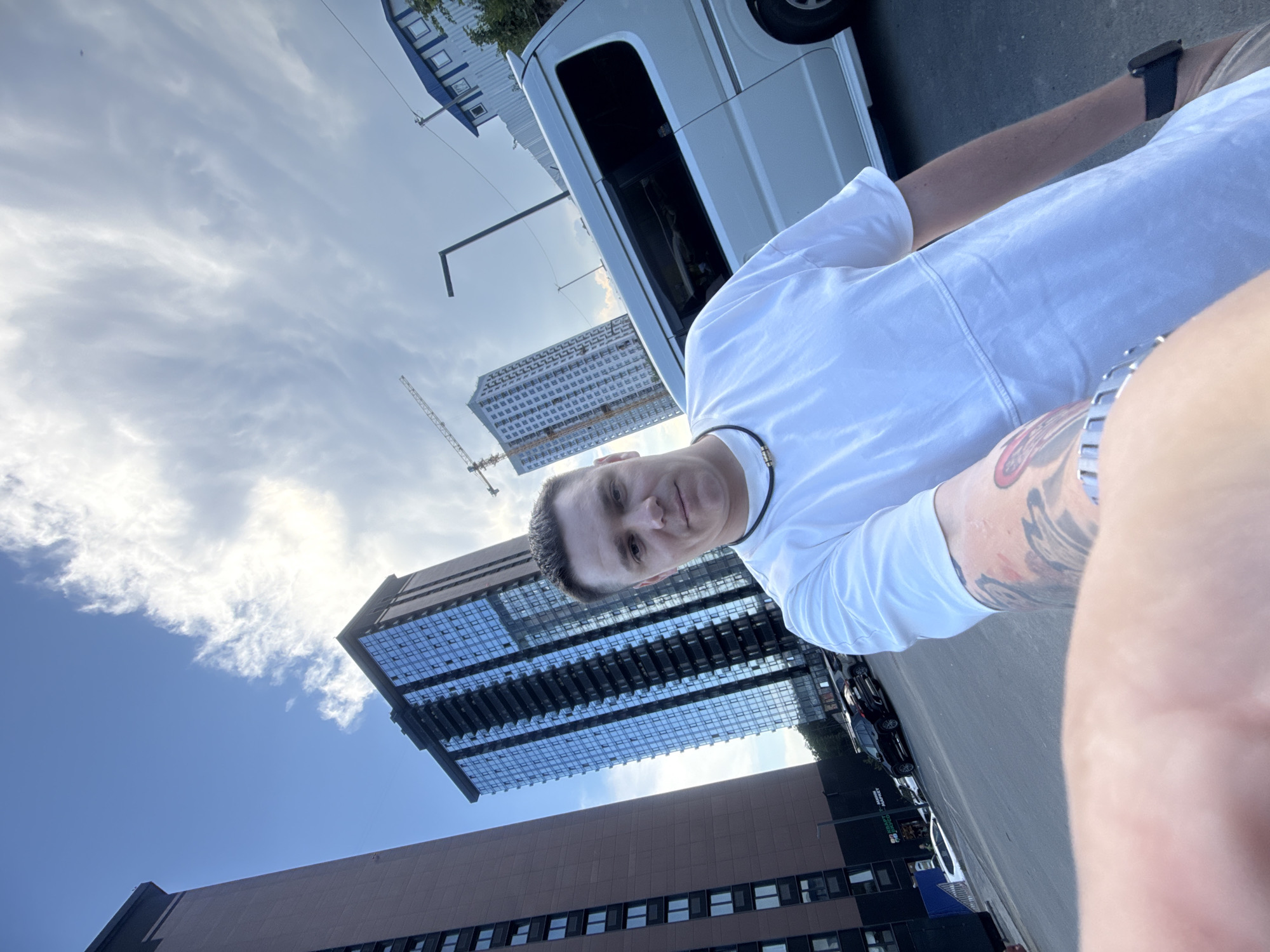 A low-angle, rotated selfie shows a man in a white t-shirt standing outside, with a van and several buildings in the background. The man has short, dark hair and is looking towards the camera with a neutral expression. He is wearing a dark necklace. On his right arm, a tattoo is visible. The background features a white van, a modern building with reflective windows, and another building with brown paneling. A construction crane is also visible in the distance, suggesting ongoing development. The sky is blue with scattered white clouds, indicating it is daytime. The scene appears to be an urban environment. There is no visible text in the image.