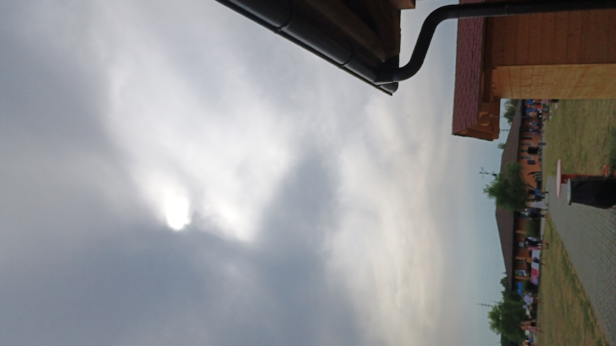 The image depicts an outdoor scene with several buildings and people, set against a cloudy sky. In the foreground, on the right side, is the corner of a wooden building with a dark gutter system. Further into the background, there is a long, single-story building with a dark brown roof, and a smaller, similarly roofed structure to its right. A paved pathway leads through a grassy area towards these buildings. Several people are visible in the middle ground, scattered across the grassy area in front of the buildings, some appearing to be walking or standing. The sky is predominantly gray with a bright, diffused spot of light breaking through the clouds, suggesting the sun is partially obscured. The overall atmosphere is overcast, typical of a cloudy day. The location is identified as Sokolov, Czechia.