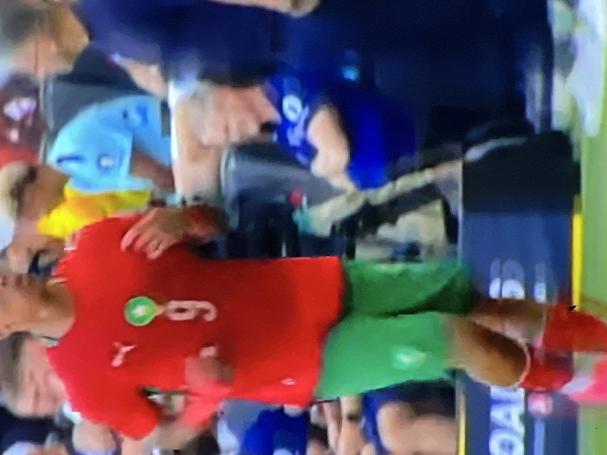 The image depicts a football player in motion on a green pitch, positioned prominently in the foreground. The player wears a red short-sleeved jersey with a white number "9" on the chest. Above the number is a green star emblem within a circular border, indicative of the Royal Moroccan Football Federation crest, and a white Puma logo is visible on the right chest area (viewer's left). The player is also wearing green shorts, red socks, and pink/red football boots. Their skin tone is of a brown complexion. In the background, to the right, a man with light-colored hair and glasses is seated, dressed in a blue short-sleeved shirt with arms crossed, next to another standing man in a dark blue shirt. Other blurred figures and spectators are visible in the far background. A blue advertising board with partially visible white text, including "OAL" and "5," runs along the lower edge of the background. The scene is set within a stadium or training ground, in the city of Casablanca, Morocco, as indicated by the player's national team attire.