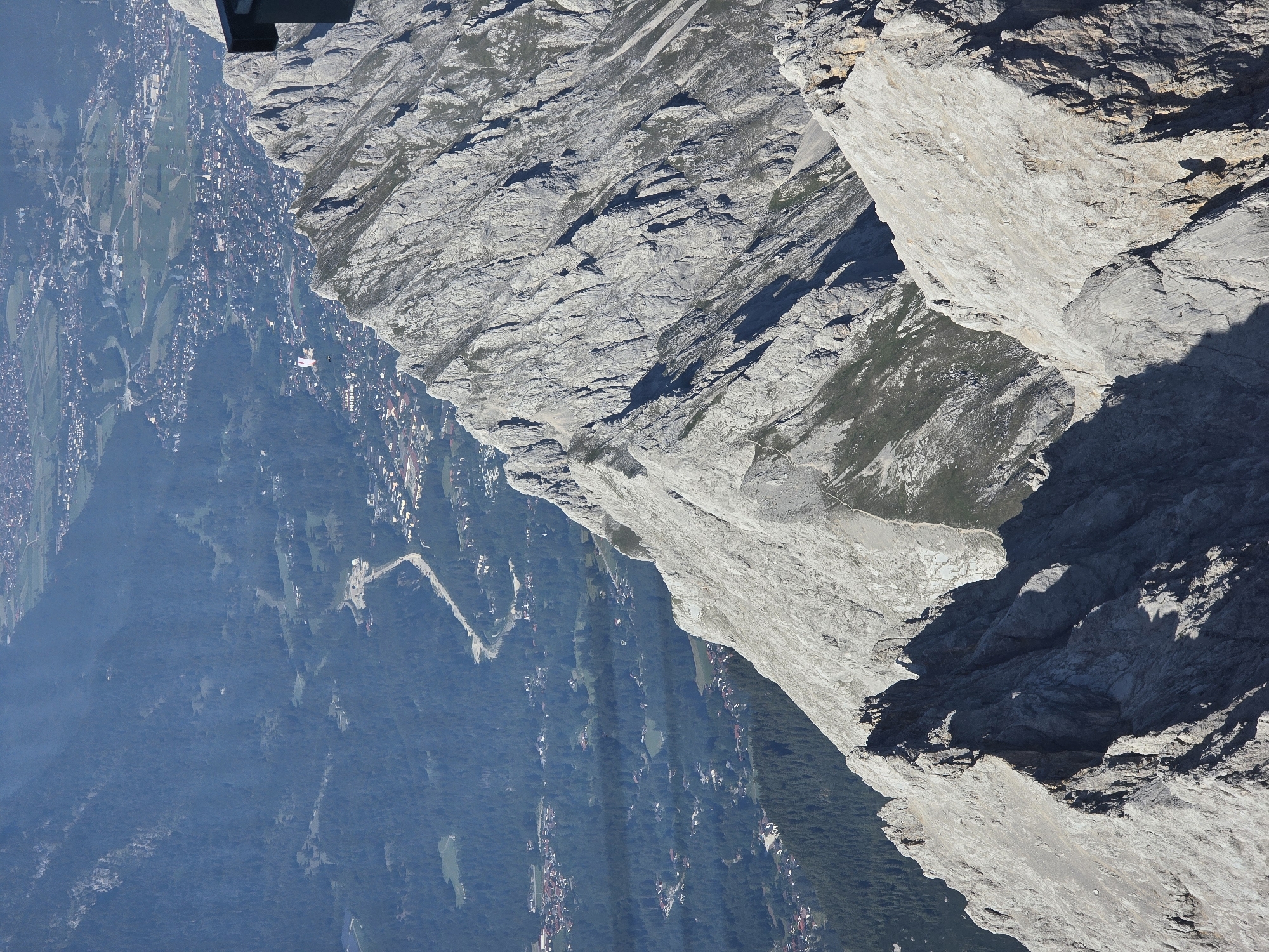 This is a high-angle, downward-facing shot of a mountainous landscape with a village nestled in the valley below. The scene appears to be taken from a very high vantage point, possibly from a cable car or a drone. The rugged, grey rock face of a mountain dominates the foreground and right side of the image, showing intricate textures and deep shadows. Below and to the left, a verdant valley unfolds, dotted with a settlement of buildings, some appearing to be houses and others larger structures. Further into the valley, dense green forests cover the slopes, interspersed with patches of lighter green fields. A winding road is visible, leading through the settlement and into the forest. There's a hint of a paraglider, a small white shape with a colored wing, suspended in the air above the valley, suggesting recreational activity. The lighting indicates it's daytime, likely late morning or early afternoon, with strong sunlight casting distinct shadows. The weather appears clear and sunny, ideal for aerial views and outdoor activities. The location is Garmisch-Partenkirchen, Germany, which is known for its dramatic alpine scenery and outdoor pursuits.