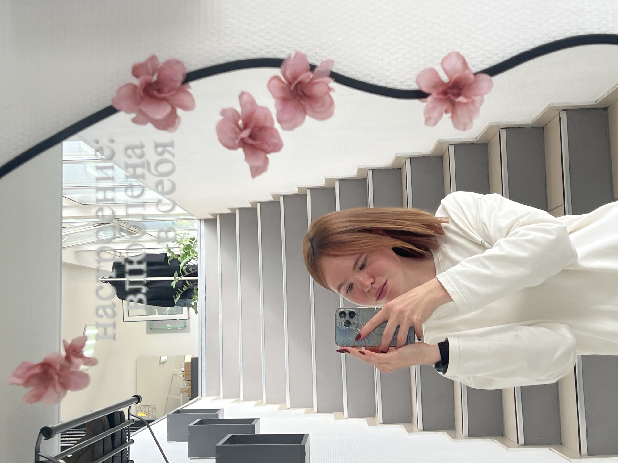 A young woman with shoulder-length auburn hair takes a selfie in a mirror. She is wearing a long-sleeved white shirt. She holds a smartphone in her right hand, angled to capture her reflection. Her left hand is partially visible, holding the phone. In the background, a staircase with gray and white steps ascends. The mirror's reflection shows more of the staircase and a black coat on a mannequin in a store. Text is visible on the mirror, appearing to be in Russian and reading "Настроение: Влюблена в себя" which translates to "Mood: In love with myself." Pink artificial flowers are attached to the mirror's frame, with some reflected in the glass. The scene appears to be indoors, possibly in a retail or public space. The location is identified as Chaikovsky, Russia.