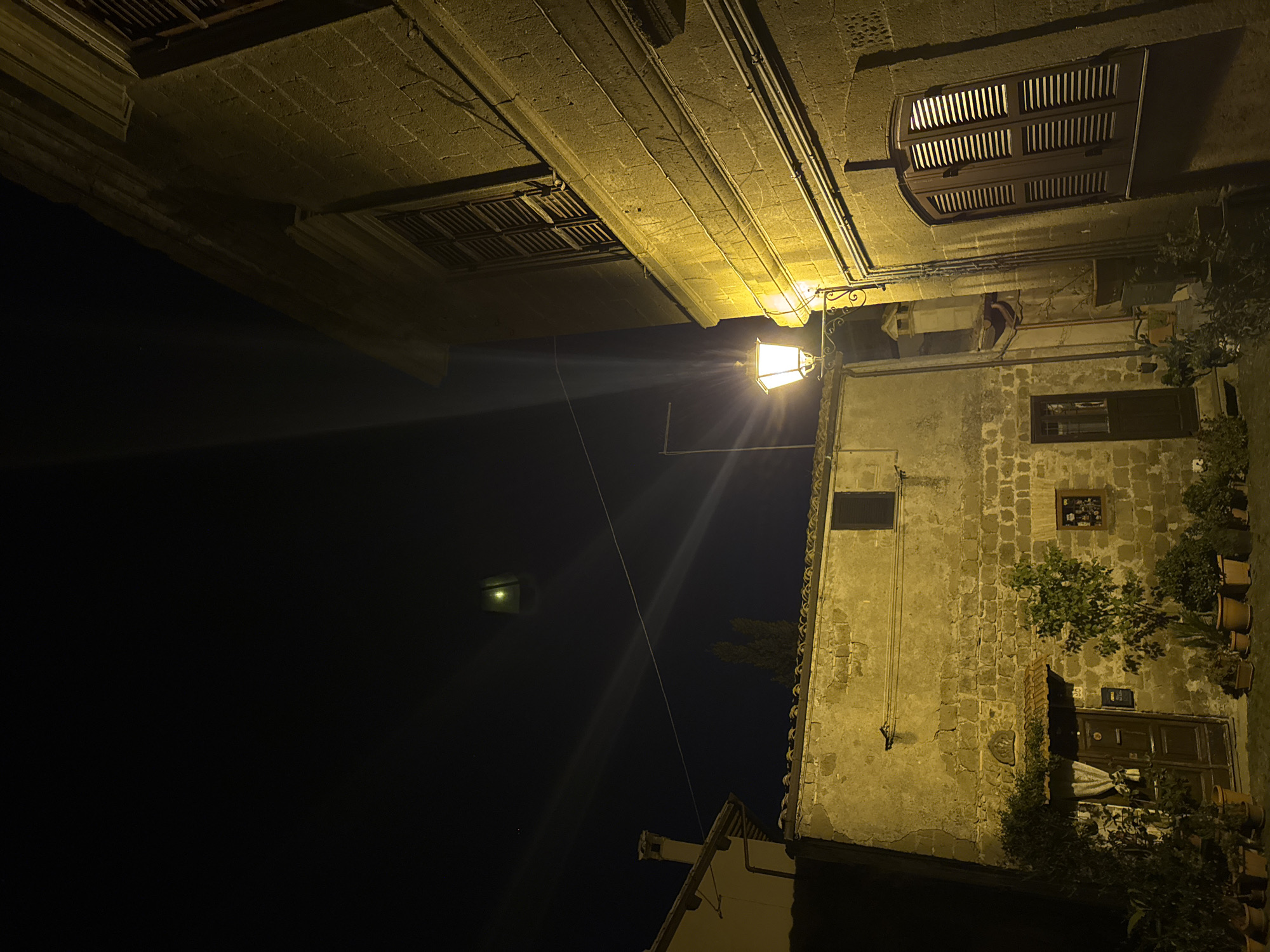 The image, oriented vertically, presents a night scene in Castelnuovo di Porto, Italy. A street lamp, illuminated and emitting beams of light, is affixed to the side of a multi-story stone building. The building, exhibiting signs of age and wear, features a wooden door, windows with shutters, and potted plants lining the entrance. Above, a portion of another building is visible, characterized by its stone construction and window designs. The sky, dark and mostly obscured, contains a faint light in the distance. A wire stretches across the scene.