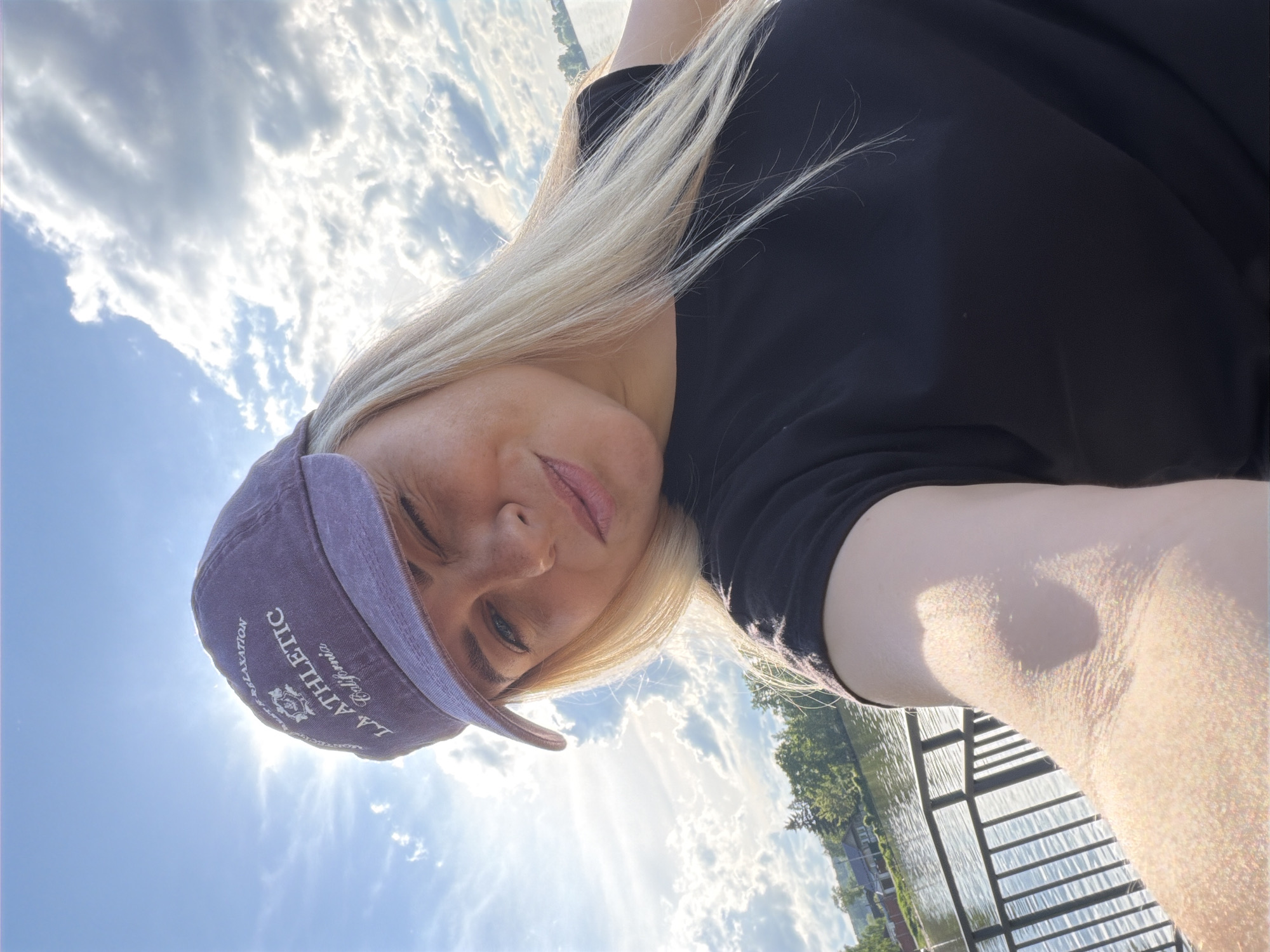 A woman with blonde hair and wearing a purple baseball cap is taking a selfie. She is wearing a black shirt. The background consists of a blue sky with puffy white clouds, a body of water, and some green trees in the distance. She appears to be standing near a metal railing or fence. The baseball cap has the words "L.A. ATHLETIC" and "California COLLECTION" written on it.
There is no information in the image to identify this location to be Votkinsk, Russia.