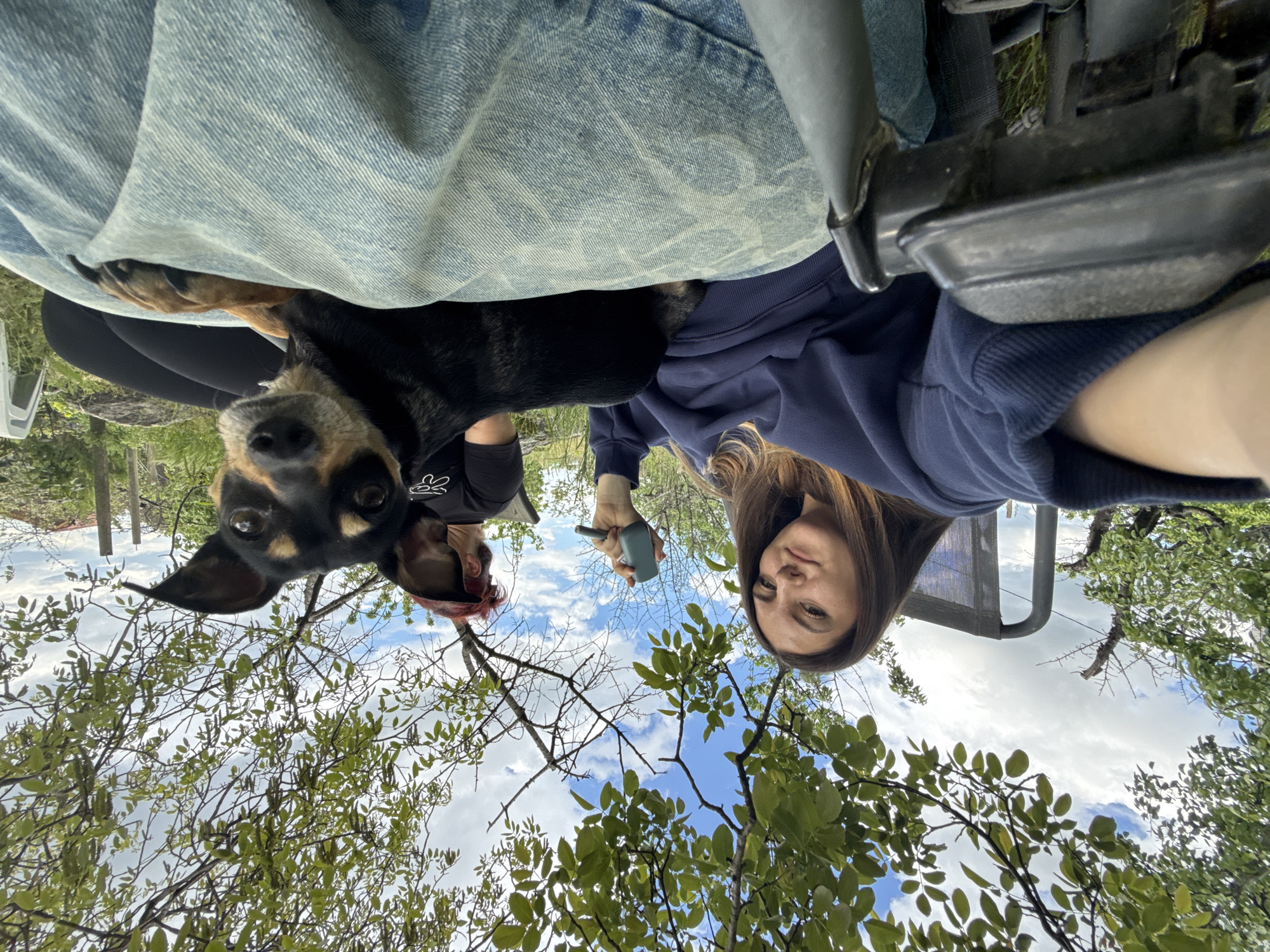 A selfie taken outdoors in daylight, with a ground-level perspective looking upwards. A woman with long brown hair, wearing a navy blue sweatshirt, holds a teal-colored object in her hand. To the left of the woman, another person with red hair and a black shirt is visible. A black and tan dog is positioned to the left of the woman. The background consists of a blue sky with white clouds and green trees. A denim-like fabric and part of a black chair frame are also visible. The location cannot be specifically determined to be in Sliven, Bulgaria based on visual cues alone.