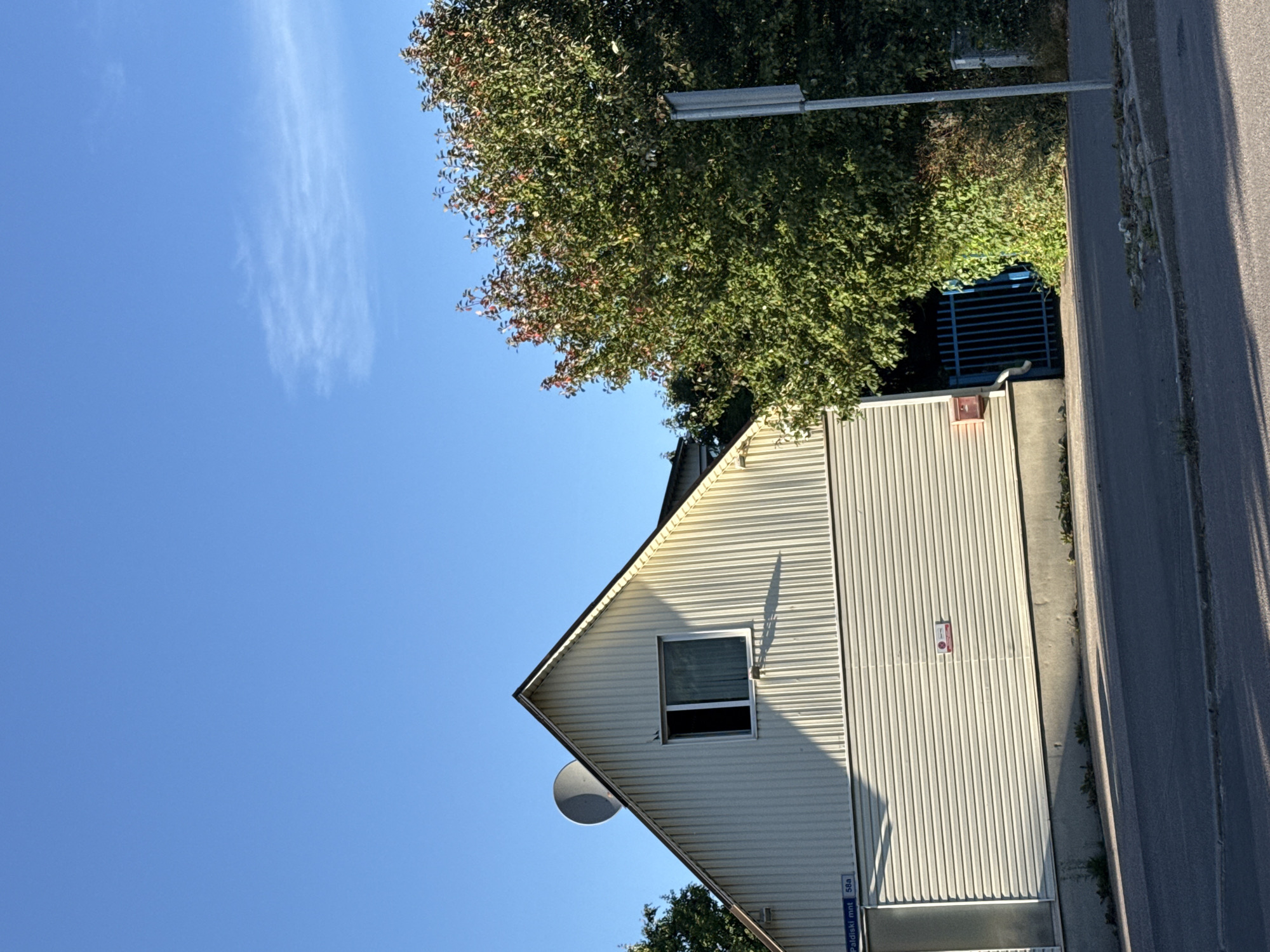 The image shows a portion of a light-colored, corrugated building with a gable roof. A satellite dish is mounted on the left side of the gable, above a single window. The building's lower section is also covered in the same corrugated material. To the right of the building, a dense green tree with some red foliage is visible. In front of the tree and partially obscured by it, a blue metal gate is present. A gray pole with a sign is located in the foreground to the right. The sky is clear and blue. A street and sidewalk are in the foreground. Street signage is visible on the building indicating "Paldiski mnt 58a". This location is on Paldiski Highway in Tallinn, Estonia.
