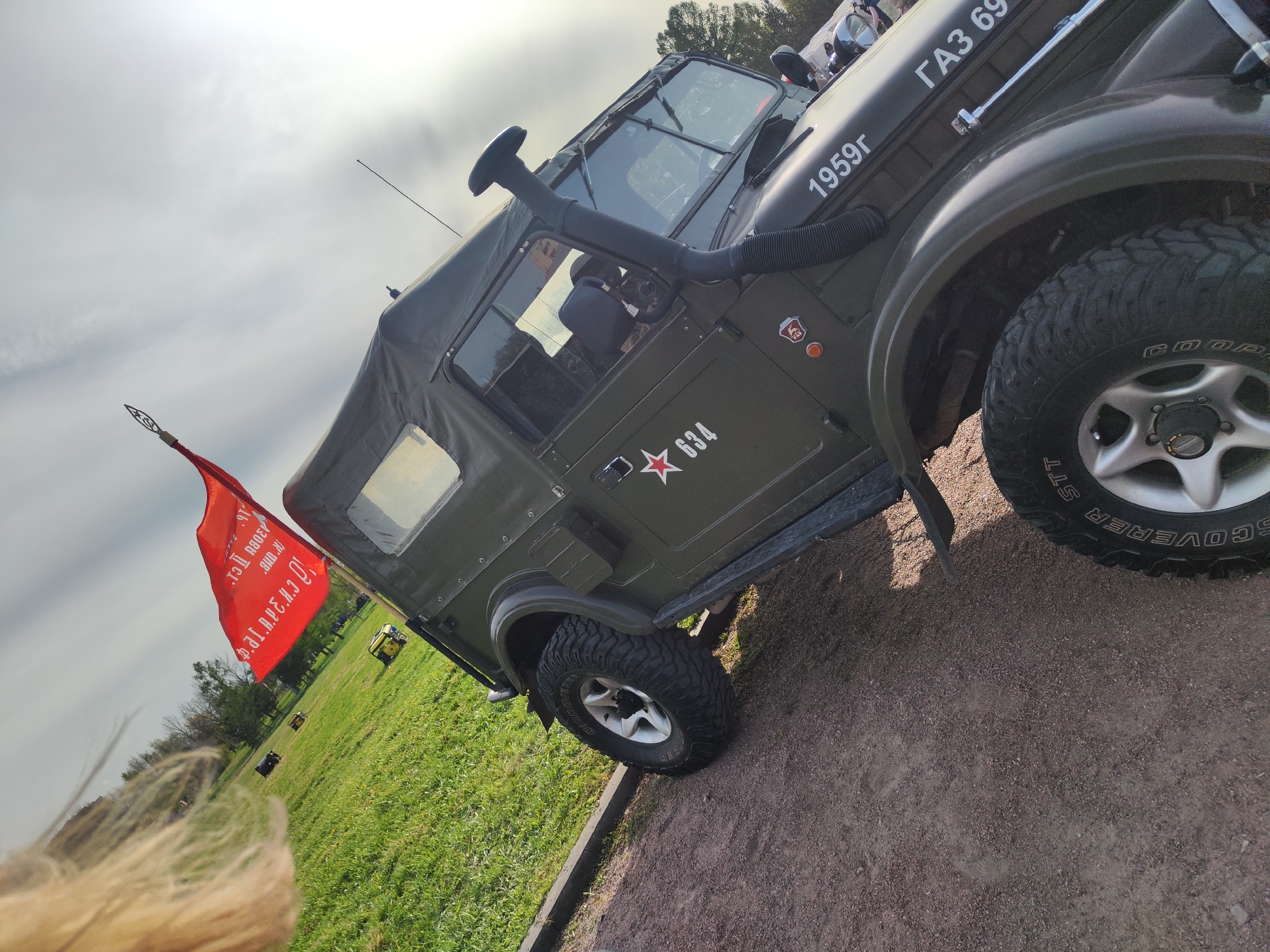 The image shows a GAZ-69 Soviet four-wheel drive light truck, likely at an outdoor event in Unknown, Unknown. The truck is painted in an olive-green color. It is parked on a patch of gravel or dirt, next to a grassy area. The truck has a snorkel intake and a red flag attached to its rear. The flag has text written in Cyrillic. The year "1959г" is painted on the truck's hood, along with the letters "ГАЗ 69". A five-pointed red star and the number "634" are stenciled on the truck's side. In the background, there are other vehicles and people.