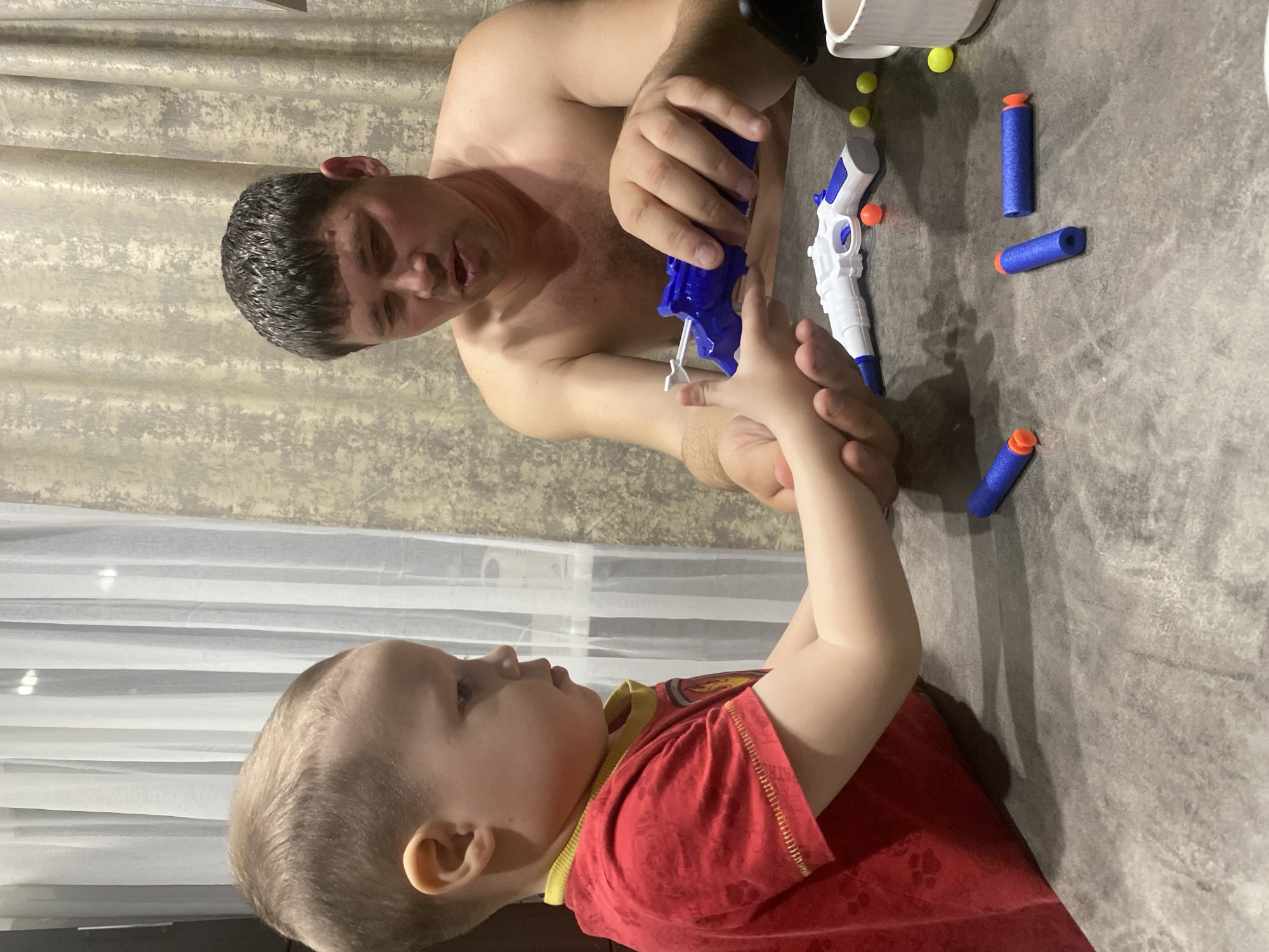The image captures an intimate indoor scene featuring an adult male and a young child engaged in playful interaction with toy guns. The photo is taken from a slightly elevated perspective, looking down on the two individuals who are positioned on a grey, textured surface, possibly a floor or a large low table.
On the left side of the frame, an adult male is depicted from the waist up. He is shirtless, revealing a muscular build and light body hair. He has short, dark hair that appears damp. His gaze is directed towards the child, and his expression suggests focus and engagement. His hands are holding a bright blue toy gun, which resembles a water gun or a soft-dart blaster, and his fingers are wrapped around its handle.
To the right, a young child, likely a boy, is visible from the chest up, lying or sitting on the same grey surface. He has light, short hair and is wearing a red t-shirt with a yellow trim around the collar. The child is looking up intently at the adult and the toy gun. His left hand is raised, reaching towards the adult's hand or the blue toy gun, indicating active participation or curiosity in the play.
Scattered on the grey surface around the two individuals are various toy elements. There are several blue foam darts with orange tips, typical of toy blasters, as well as a few small yellow and orange spherical balls, which might be ammunition for a different type of toy. Another toy gun, white with blue accents, is resting on the surface near the adult's hand. A portion of a white cup or container is also visible in the upper right corner of the frame.
In the background, behind the adult, patterned beige and grey curtains provide a textural backdrop. Behind the child, lighter, sheer white curtains are visible, suggesting the presence of a window. The lighting in the scene appears bright and even, indicating either natural daylight or good artificial illumination. The overall impression is one of a tender moment of shared play and bonding between the adult and the child. No discernible text is visible in the image.