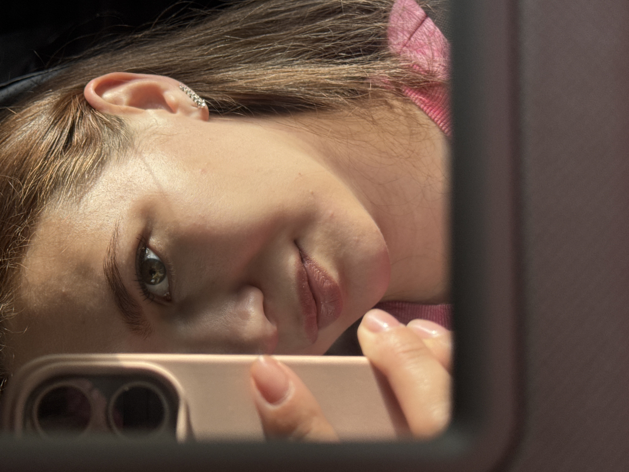 This is a close-up, sideways, selfie-style shot of a young person. They are lying down, looking into a phone that is held in their hand and used to take the photo, which is reflected in a mirror-like surface. The person has light brown hair, which is slightly tousled, and is wearing a pink top. A sparkly ear cuff is visible on their left ear. The background is dark and out of focus. The lighting suggests it is daytime, with sunlight casting shadows. The activity is taking a selfie, and the emotion conveyed is a relaxed and perhaps playful one. No text is visible in the image.