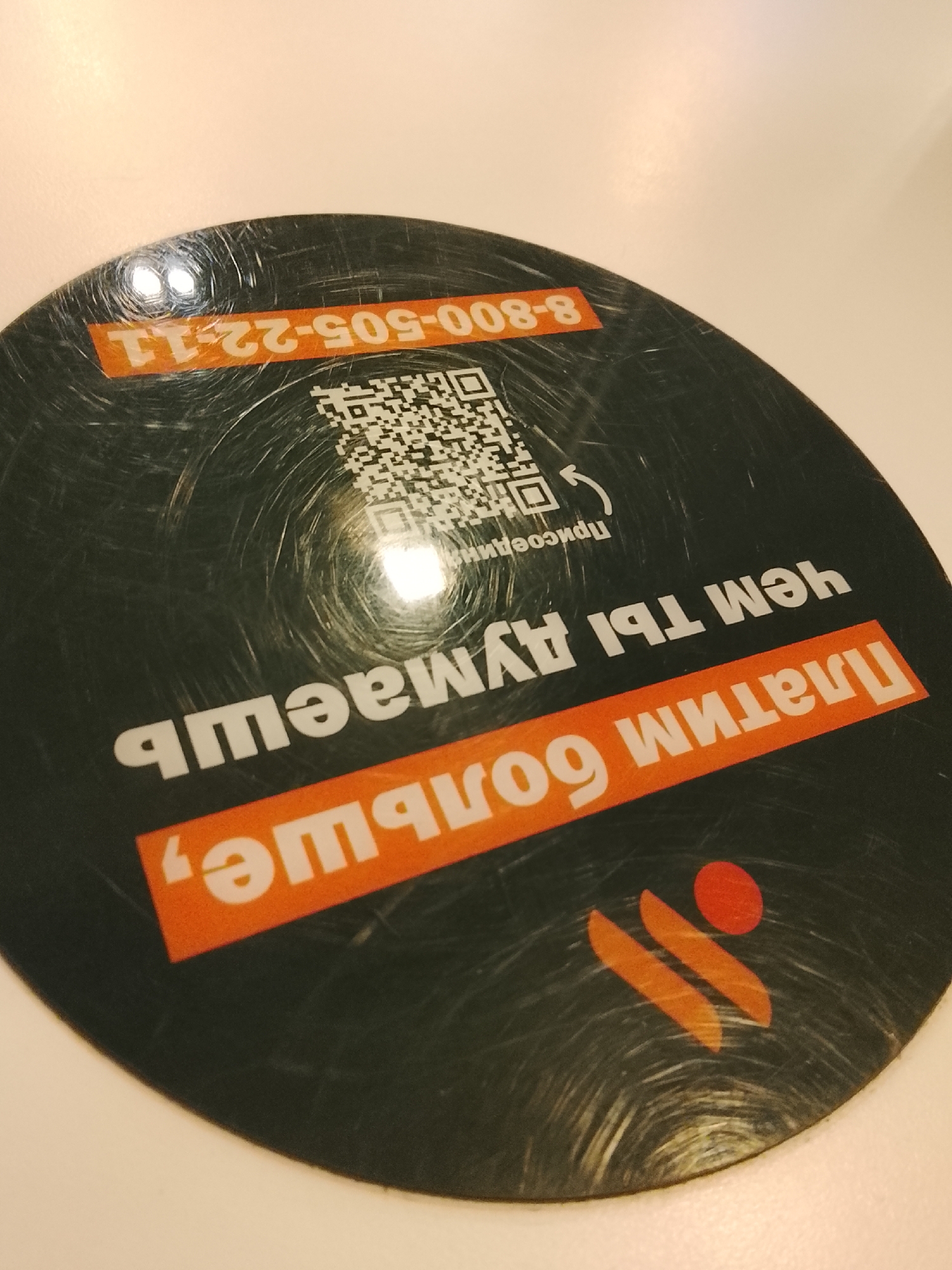 The image shows a round, black sticker on a light-colored surface. The sticker features orange text in Russian, a QR code, and a logo. The text includes the phrase "ПЛАТИМ БОЛЬШЕ, ЧЕМ ТЫ ДУМАЕШЬ" ("We pay more than you think"). The QR code is positioned near the top center of the sticker. The logo is at the bottom and appears to be of a food service company, potentially a fast-food chain. A phone number, "8-800-505-22-11," is written in orange at the top of the sticker. The sticker has visible scratches and scuffs. There is no immediate visual confirmation of the location being Obninsk, Russia, however, the presence of Russian text and a Russian phone number suggest this is the country. There are no people visible in the image.