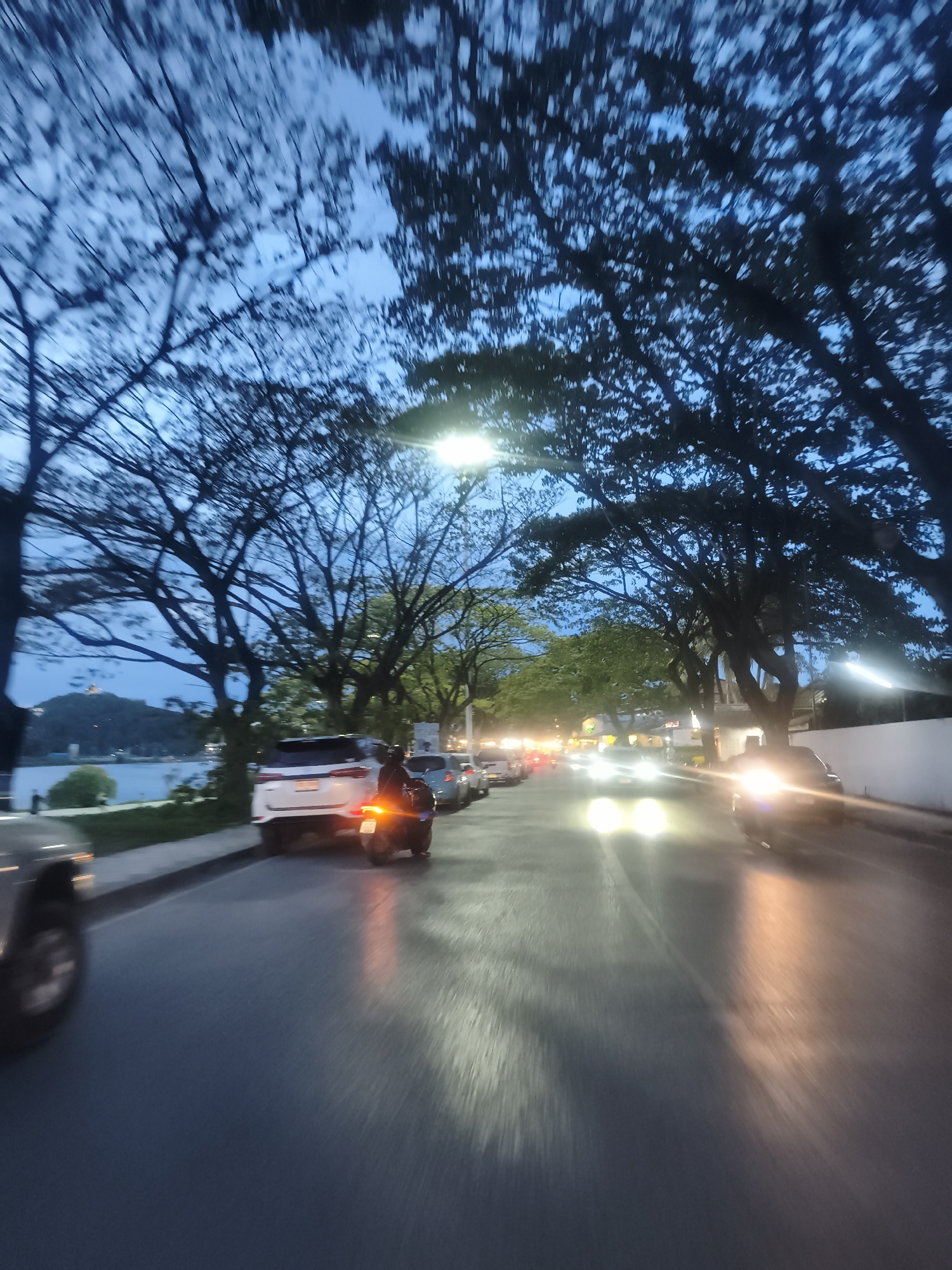 A photograph taken at twilight in Baan Chaweng, Thailand, shows a street lined with cars and motorcycles. A body of water and a distant hill are visible in the background. Trees line the street, partially obscuring the sky. The headlights of oncoming cars illuminate the road. A person on a motorcycle is stopped behind a white SUV.